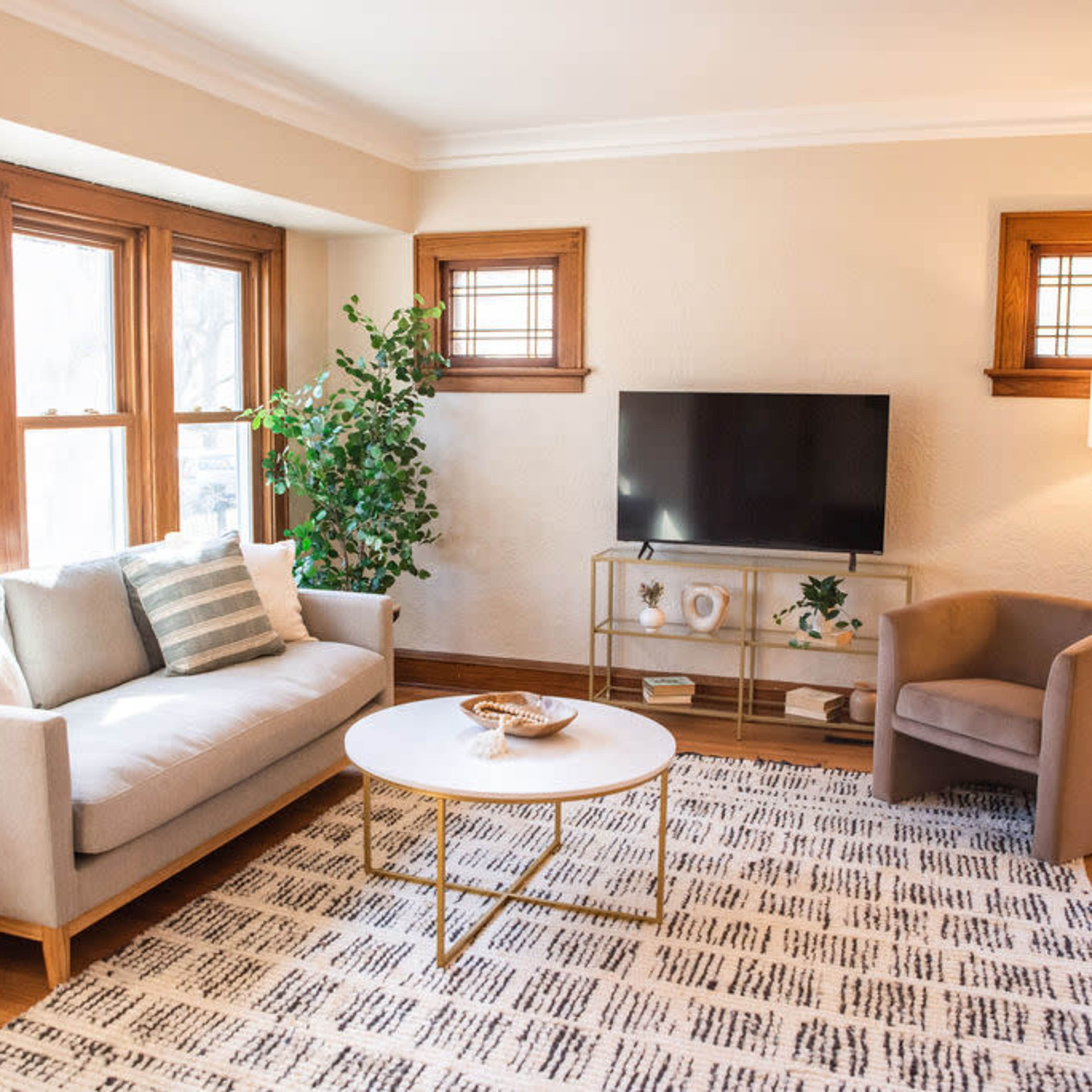 A well-lit living room featuring a beige sofa, a round coffee table, a light-colored armchair, a television mounted on the wall, and multiple windows.