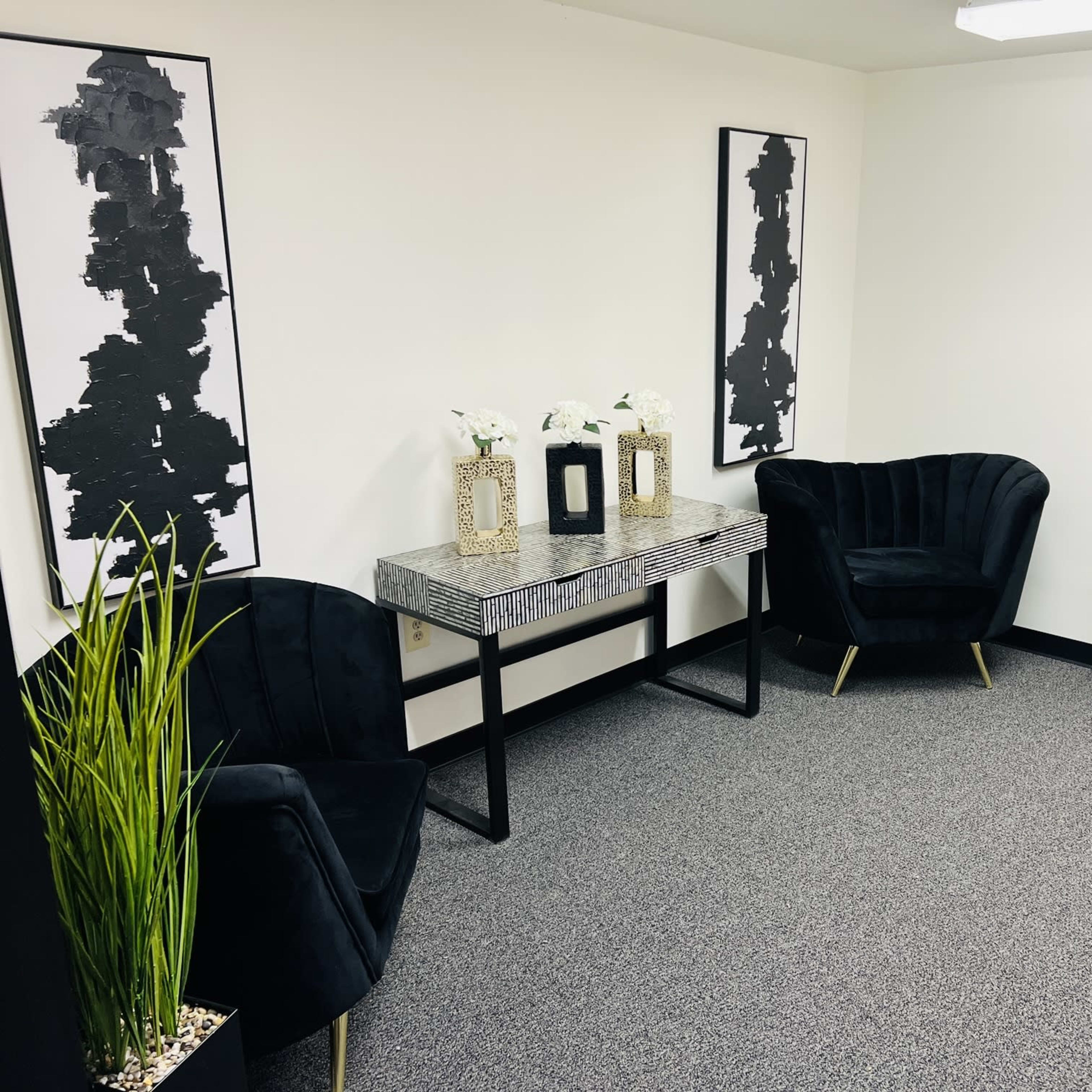 A minimalist office space with two black chairs, a patterned table, decorative plants, and framed artwork on the walls.