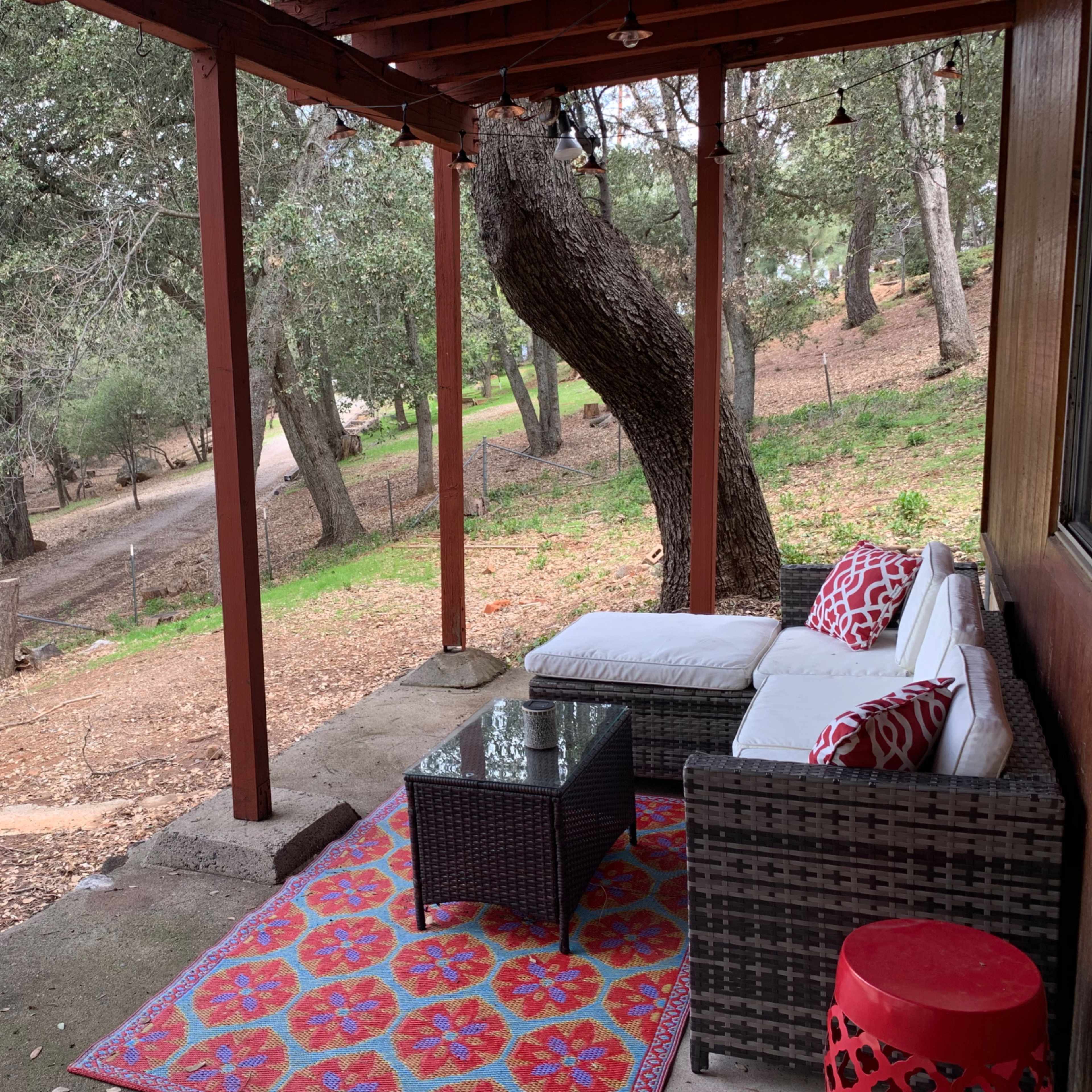 A covered porch features a seating area with a couch and glass coffee table, surrounded by trees and a gravel pathway.