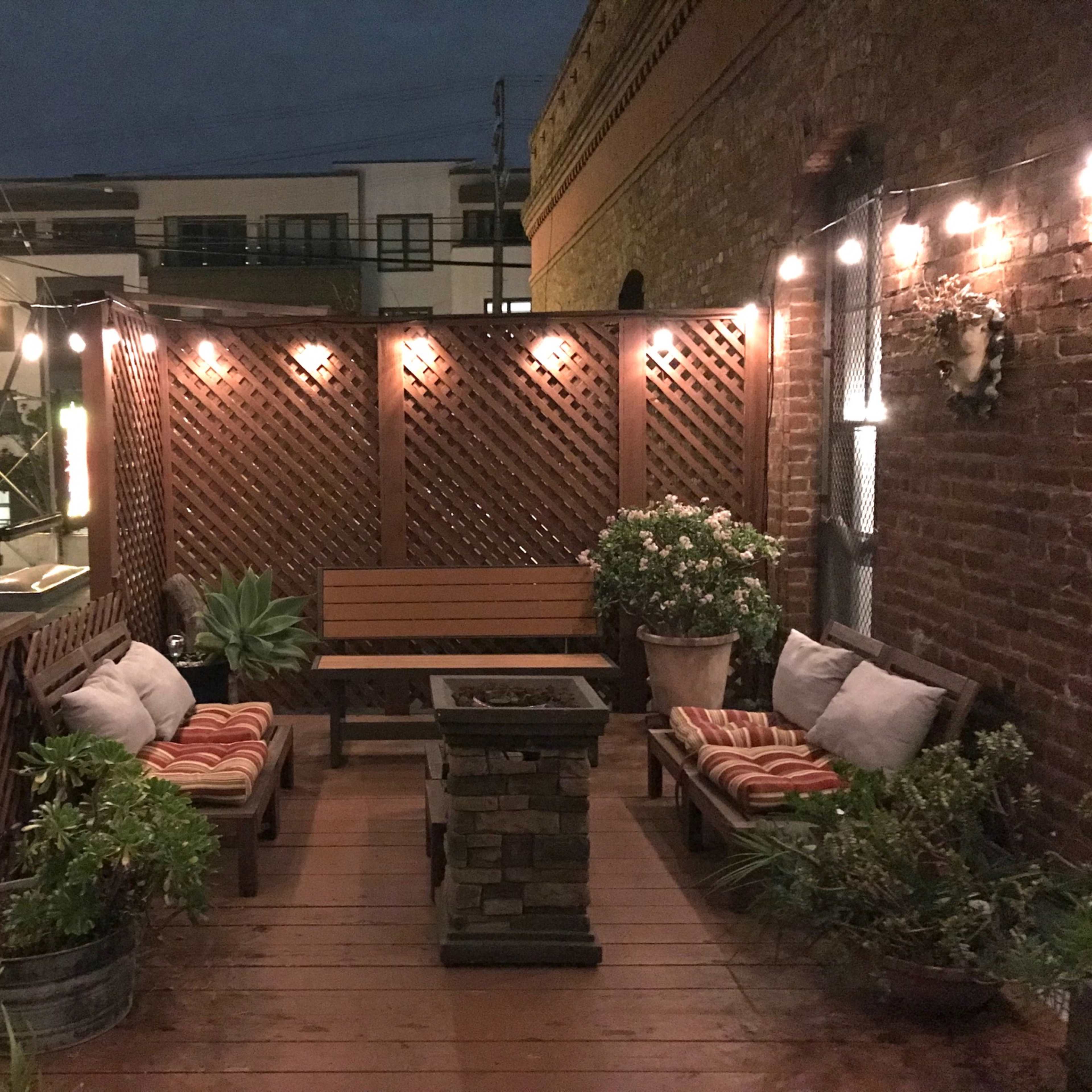 A cozy outdoor patio area with wooden seating, a stone fire pit, and string lights illuminating a lattice privacy screen.