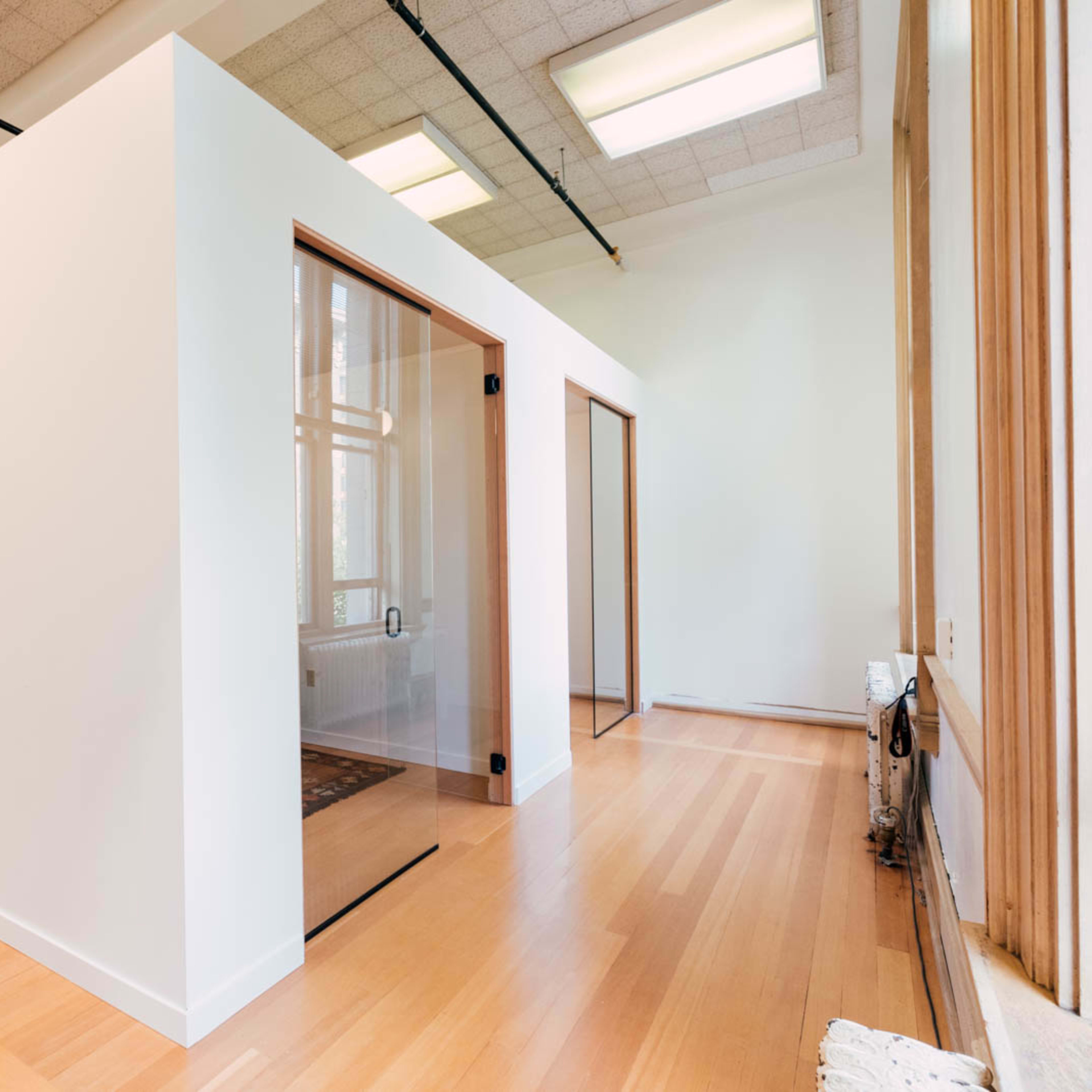 A bright, modern room with a white interior featuring light wood flooring and a glass-walled office space.