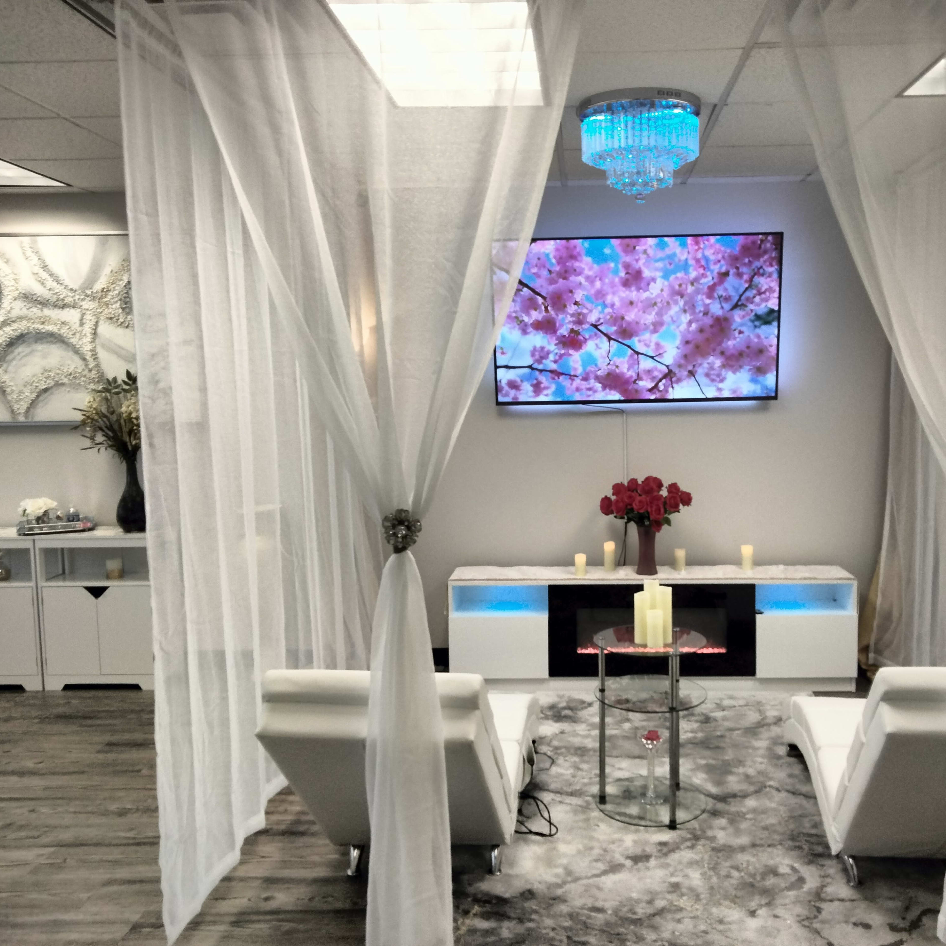 A serene lounge area featuring two white chairs, a glass table, and a large television displaying floral imagery, all surrounded by sheer white curtains.