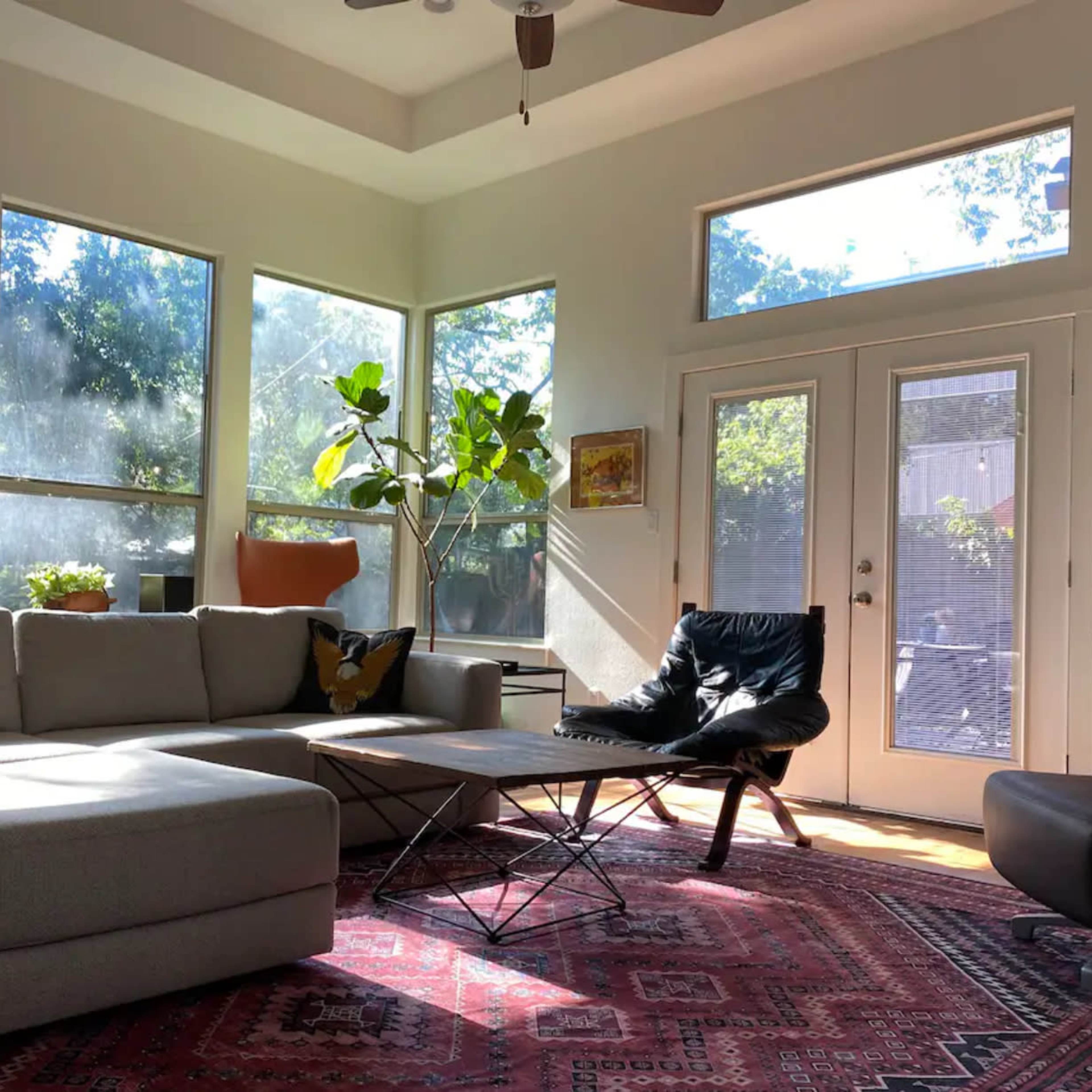 A well-lit living room with large windows, a grey sectional sofa, a black chair, and a coffee table on a patterned rug.