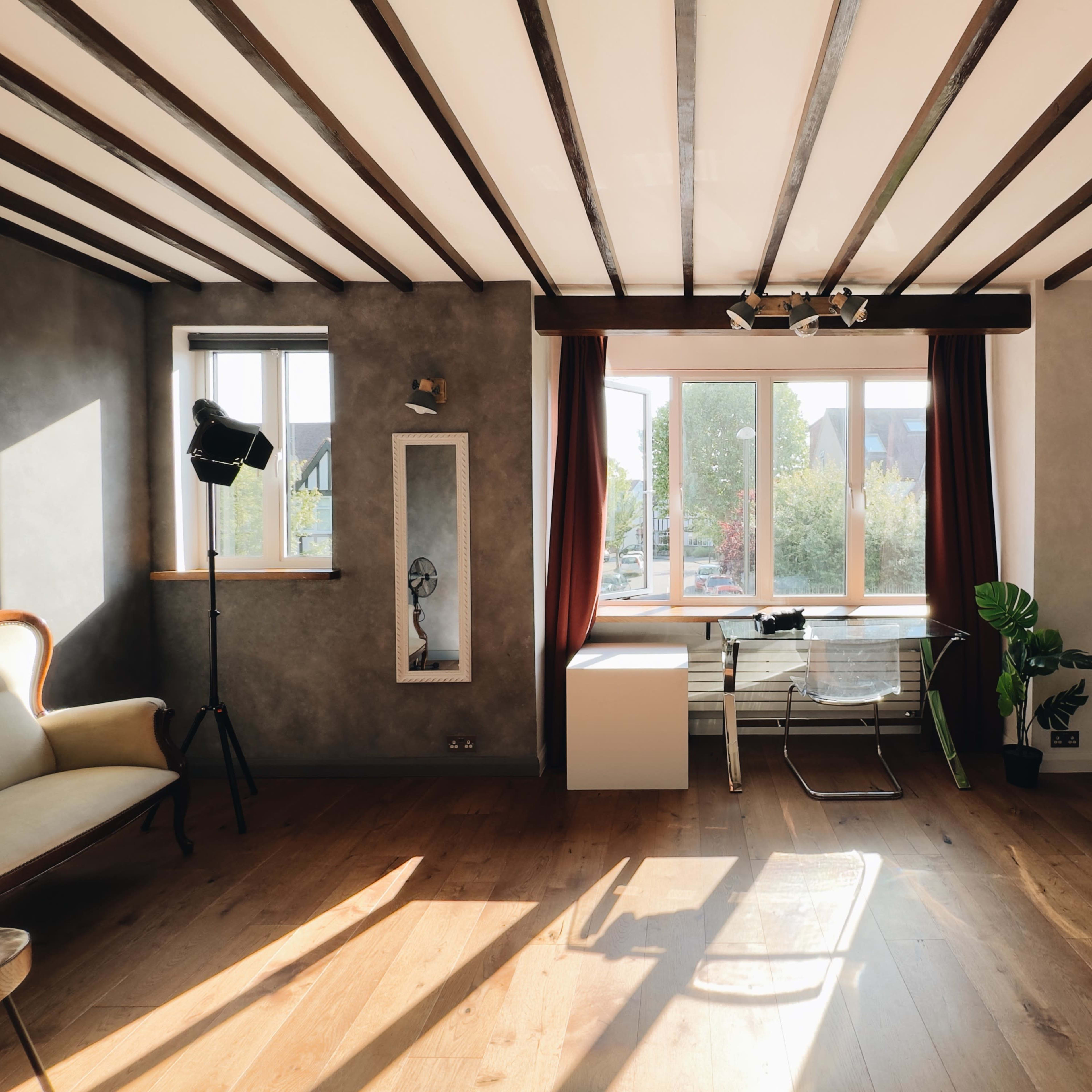 The room features wooden beams overhead, a large window with curtains, a light-colored sofa, a modern chair, and a potted plant near the corner.