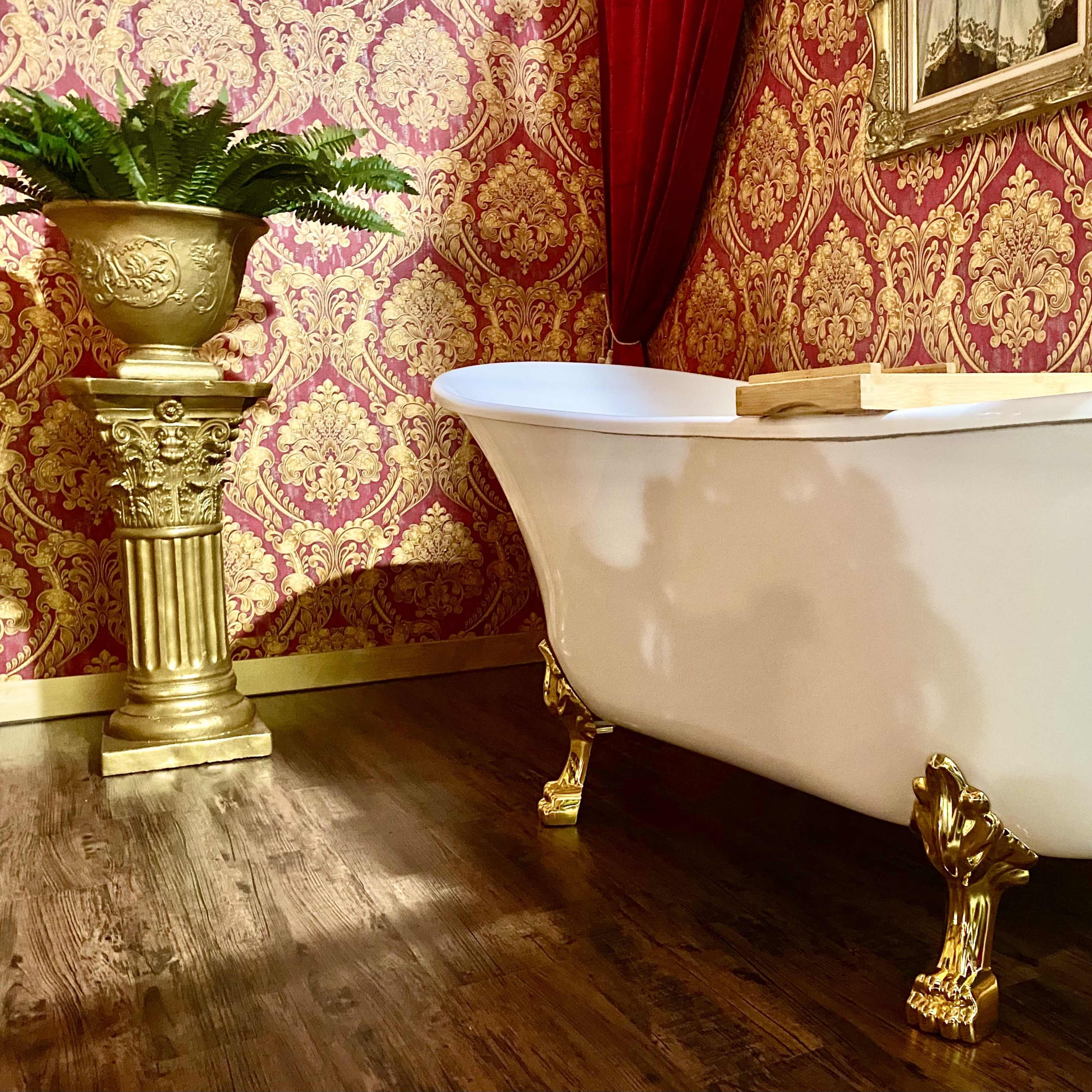 A white clawfoot bathtub with gold legs sits in a room decorated with ornate red wallpaper and a matching gold-accented plant stand.