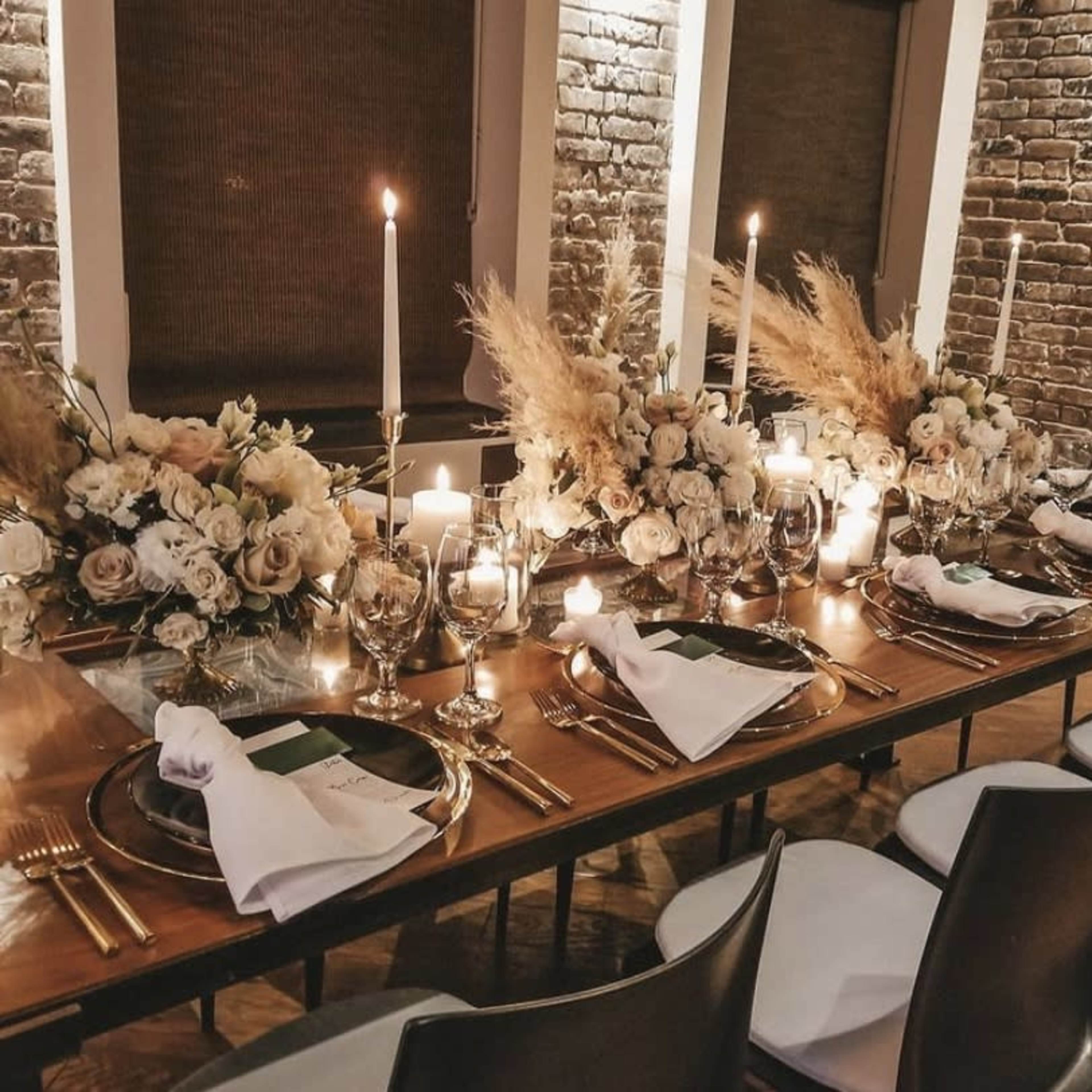 A long dining table is elegantly set with candles, floral arrangements, and glassware against a backdrop of exposed brick walls.