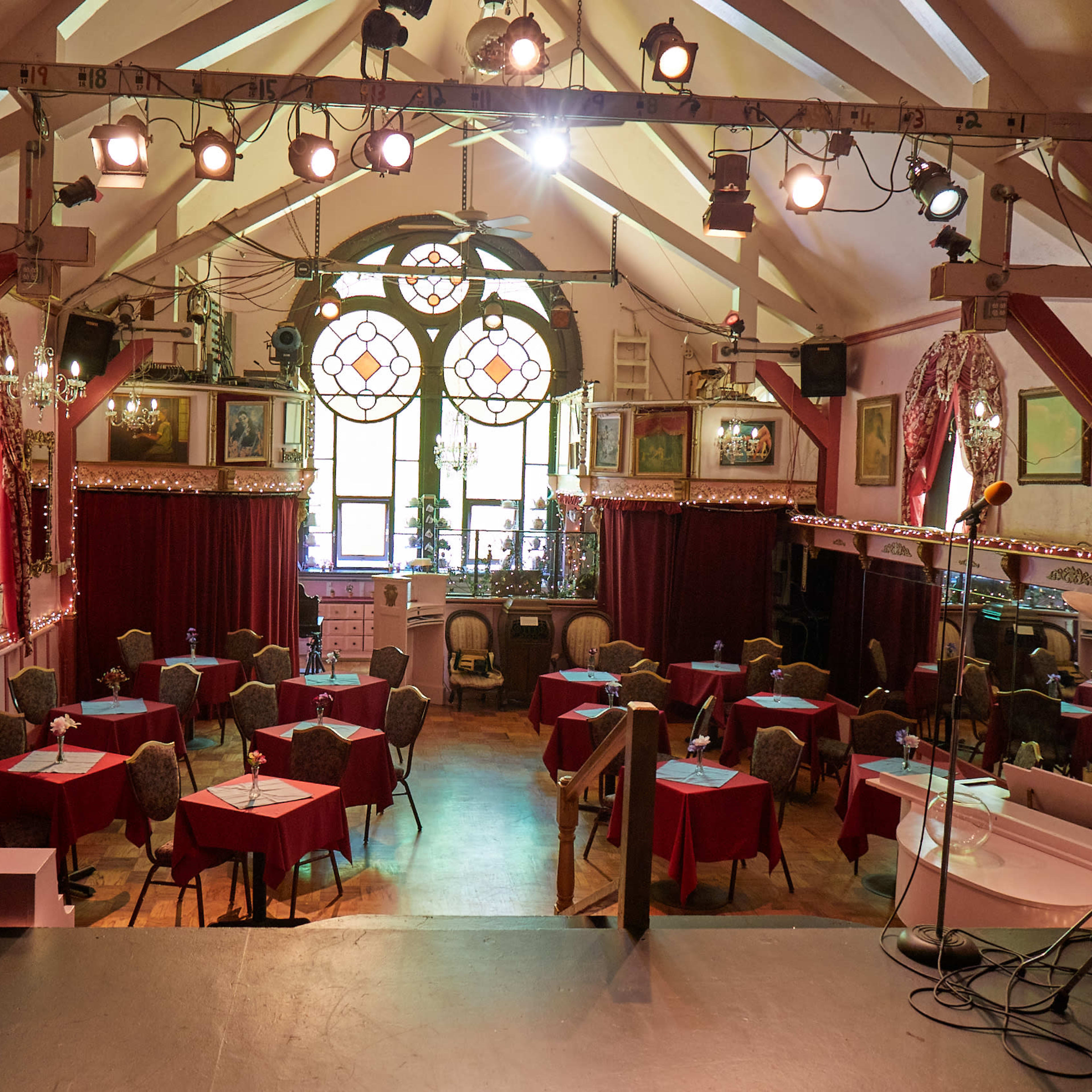 A cozy performance venue with round tables covered in red cloths, a stage in the foreground, and a large stained glass window at the back.