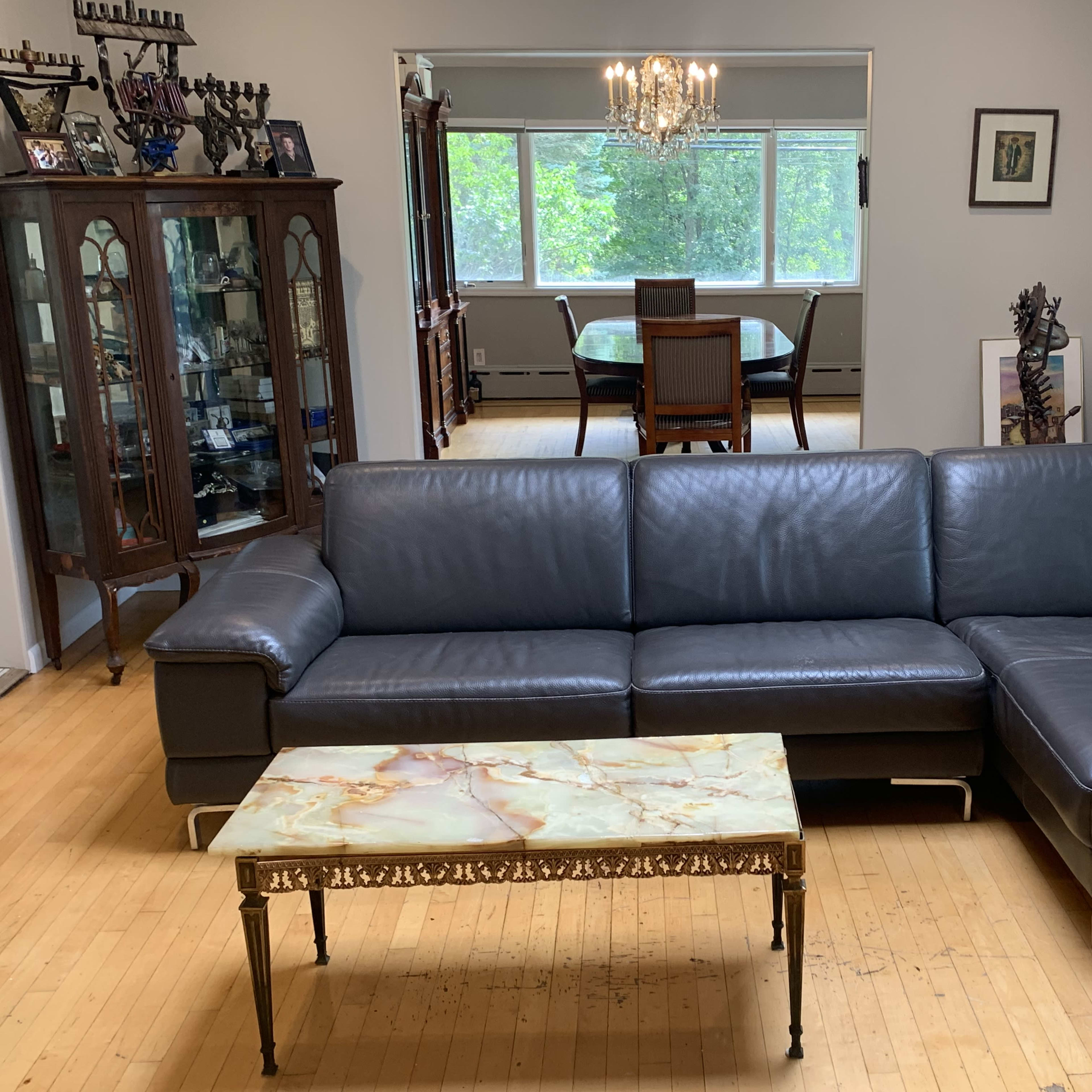 The scene features a living room with a dark leather sectional sofa, a marble-top coffee table, and a dining area visible in the background.
