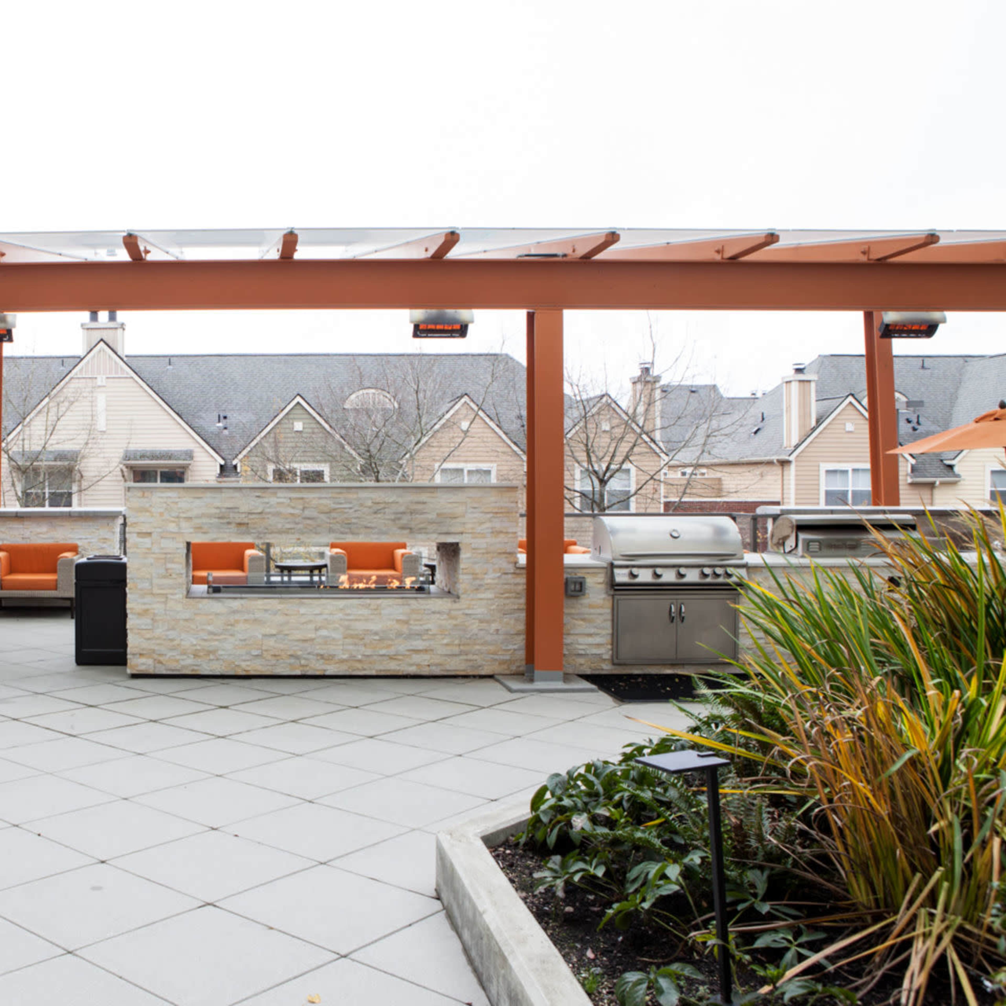 A patio area features a grill, seating arrangements under orange umbrellas, and a stone wall, with residential buildings visible in the background.