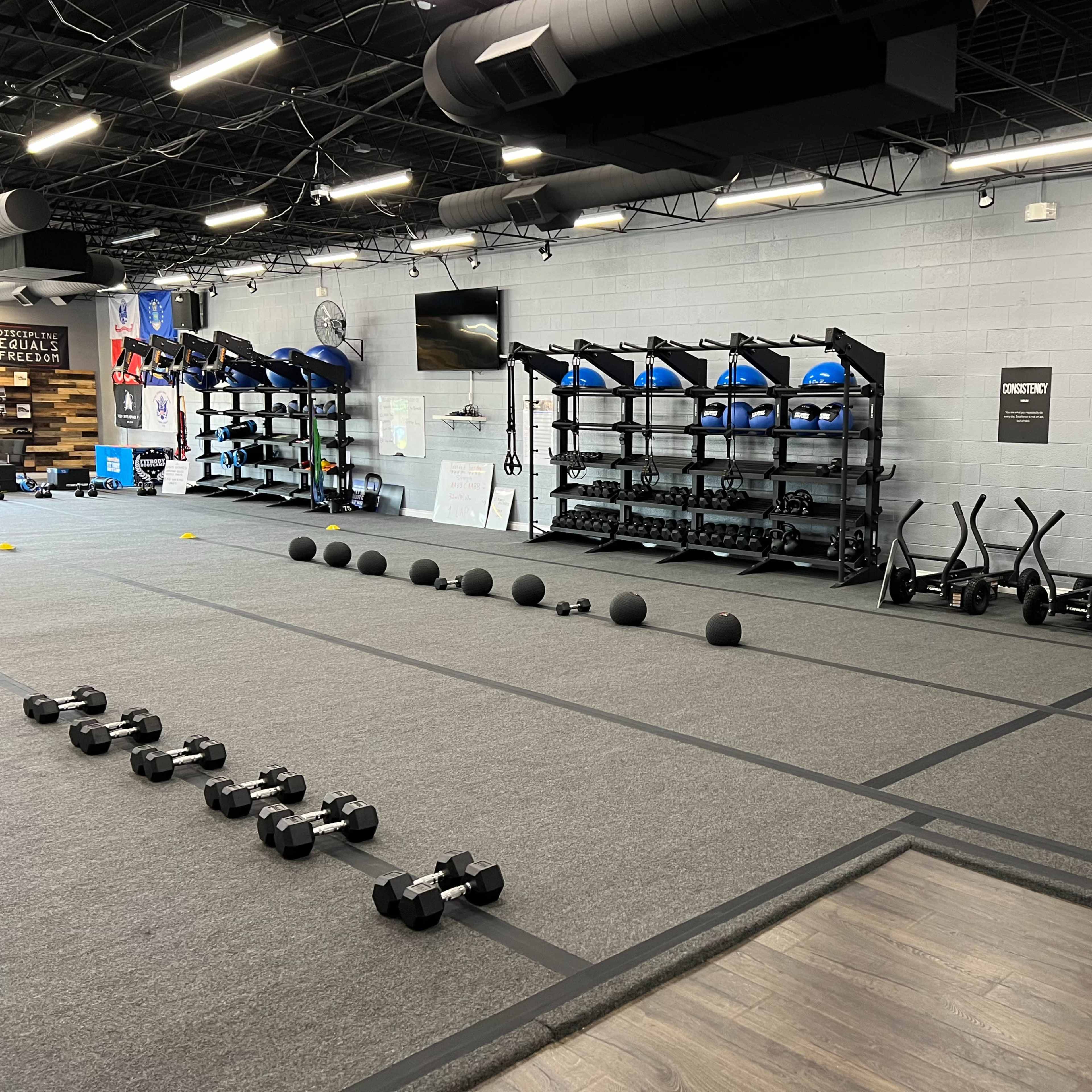 A fitness studio with various exercise equipment, including dumbbells, kettlebells, medicine balls, and stability balls organized on racks along the wall.
