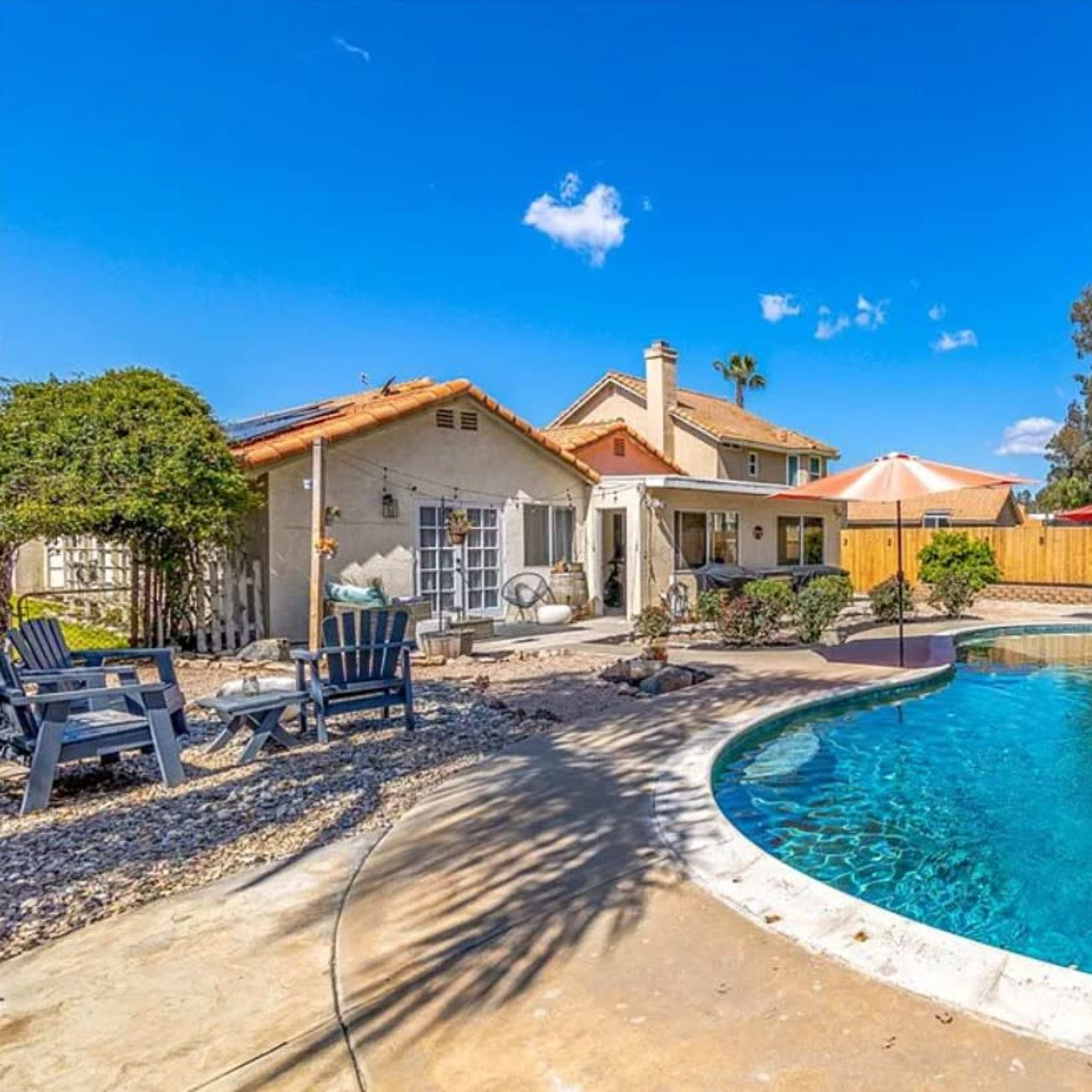 A residential backyard features a swimming pool surrounded by lounge chairs, patio umbrellas, and a house in the background set against a clear blue sky.