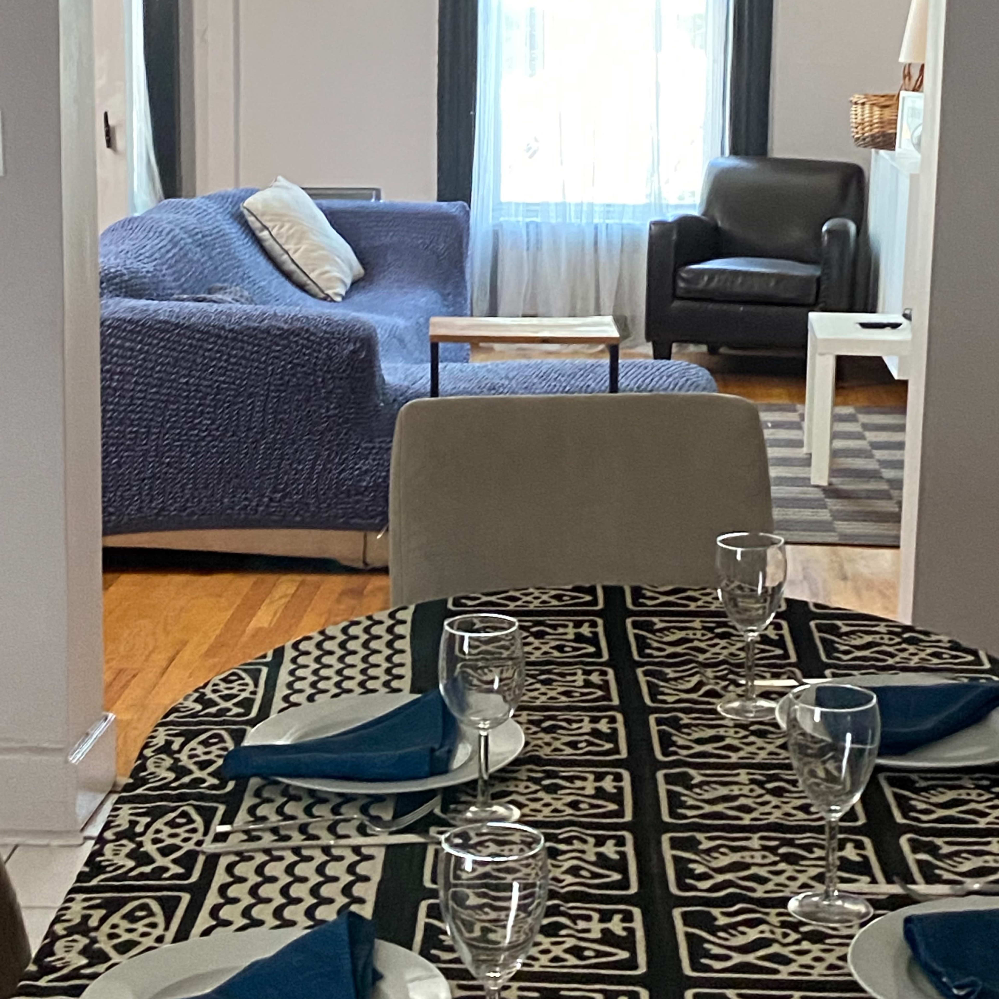 A dining table is set with glassware and blue napkins, leading into a living area featuring a blue sofa and a black chair beside a window.