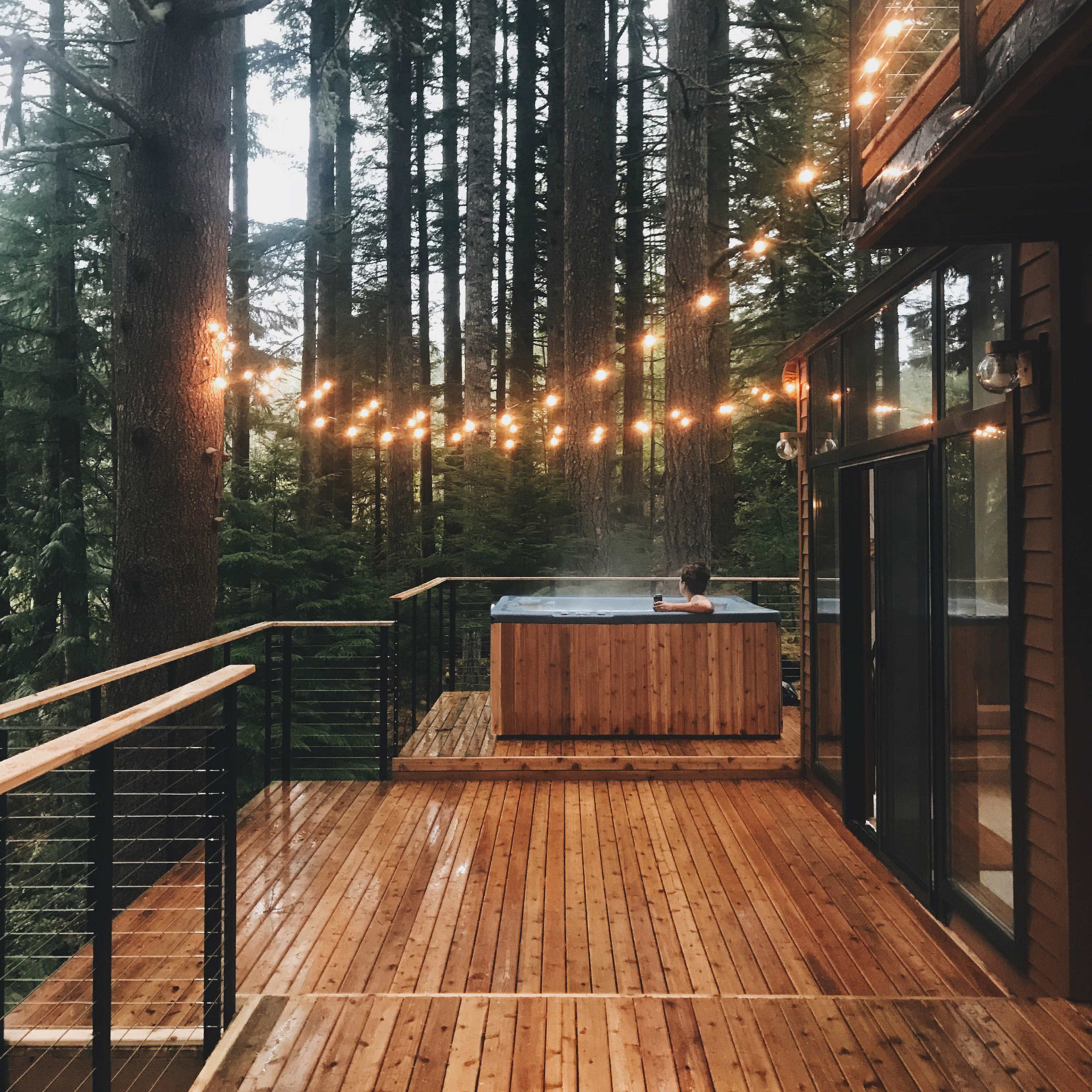 A wooden deck with a hot tub is illuminated by string lights among tall trees.