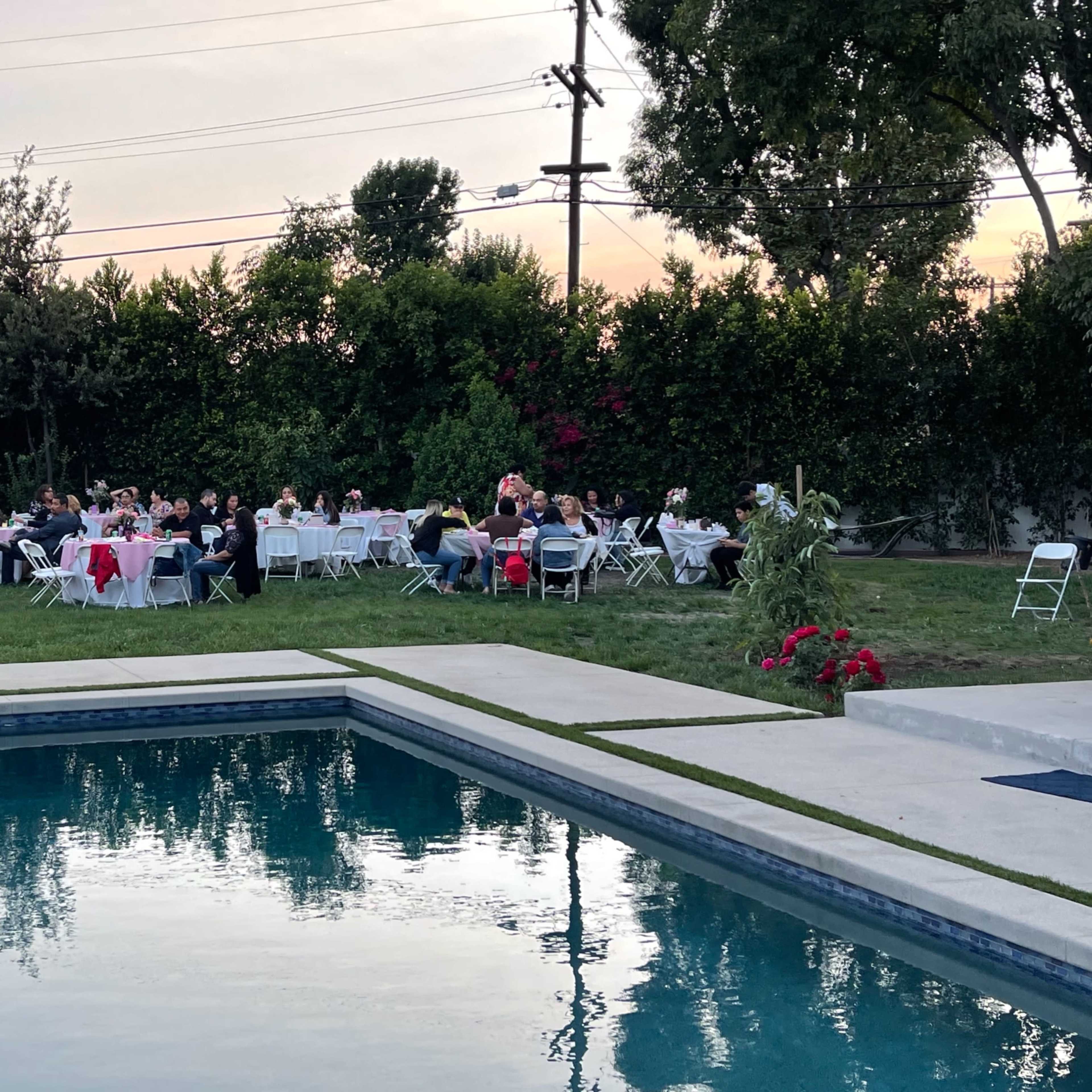 A group of people sits around tables set up for an outdoor event beside a swimming pool at dusk.