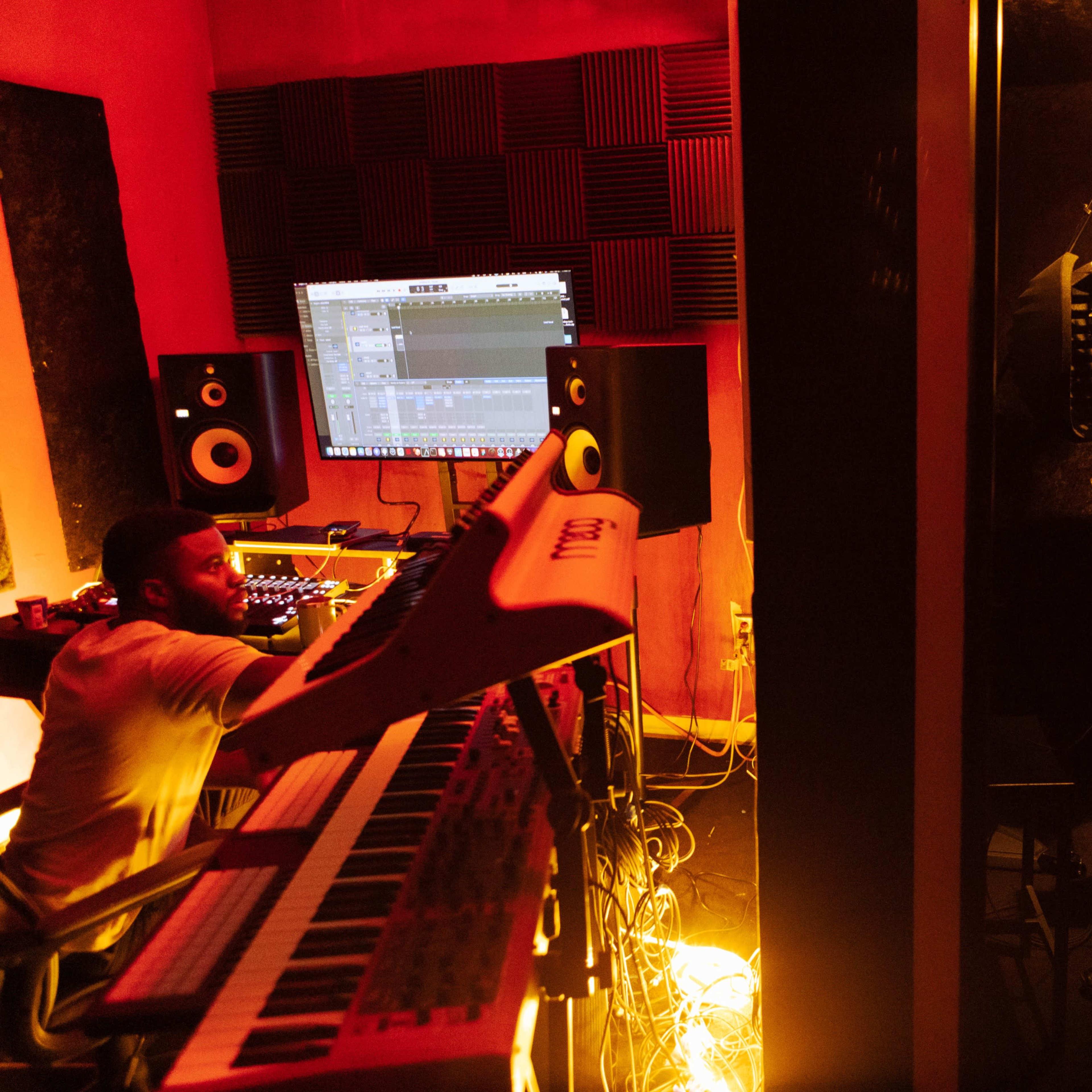 A musician stands in a soundproof booth recording vocals while another musician plays a synthesizer in a dimly lit studio.