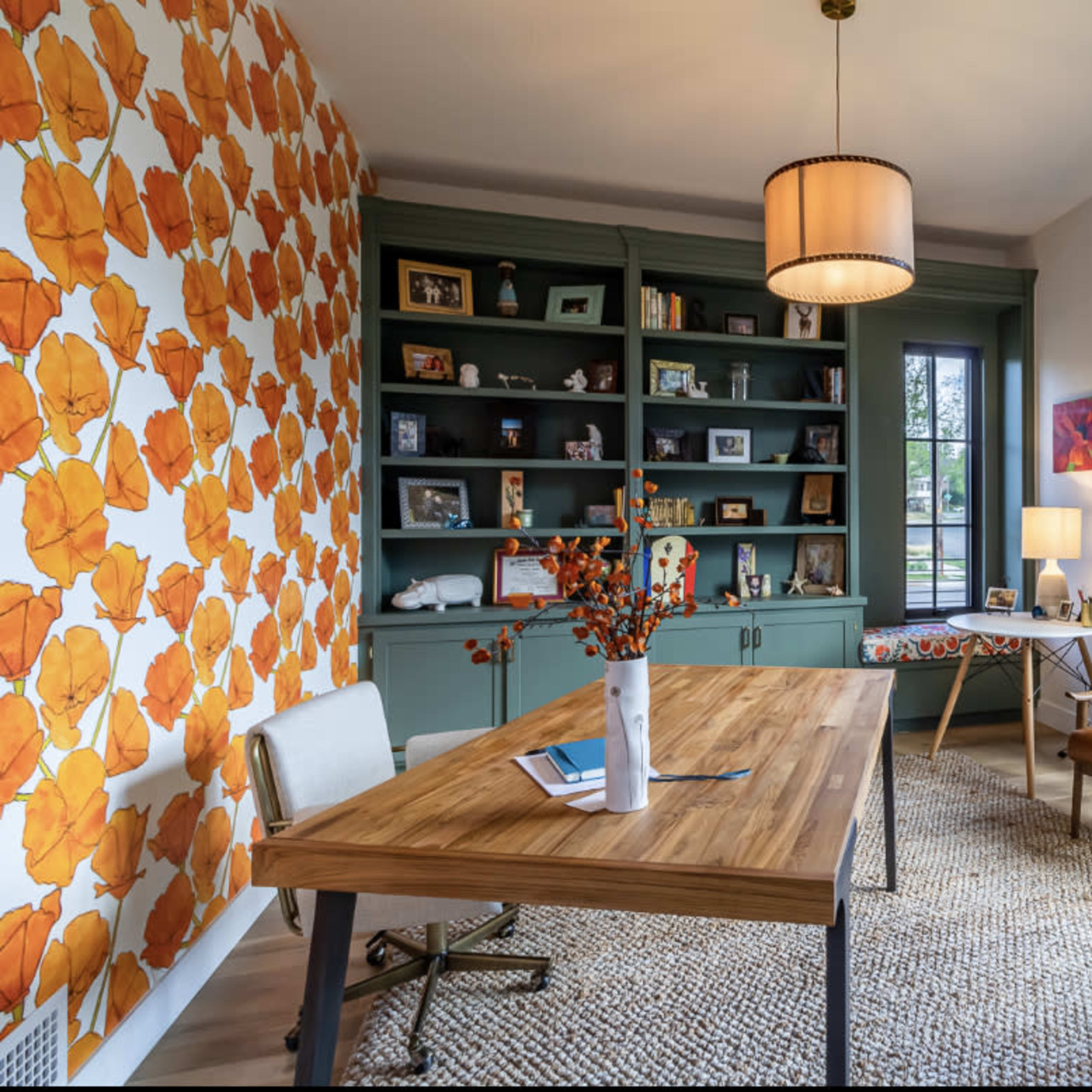 A bright room with a wooden desk, two chairs, a bookshelf filled with photos, and large windows featuring floral wallpaper and a rug.