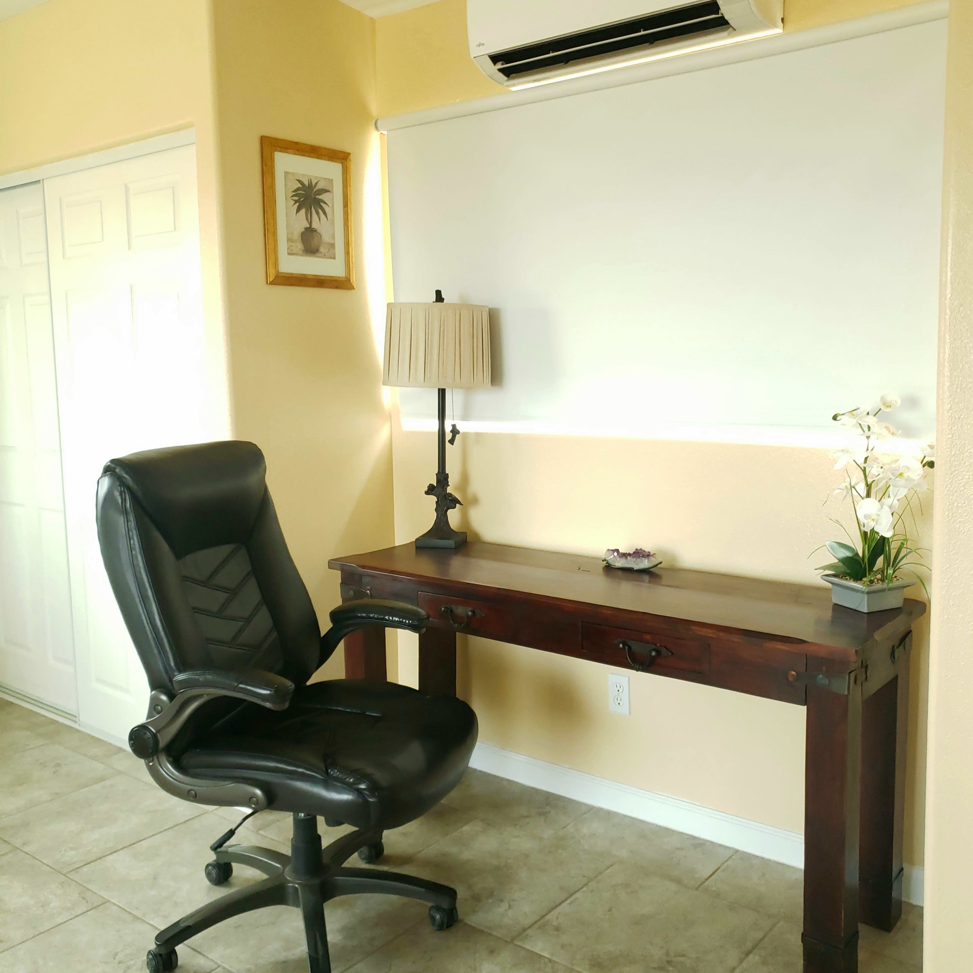 A black ergonomic chair is positioned in front of a wooden desk against a light-colored wall with an air conditioning unit and a lamp above it.