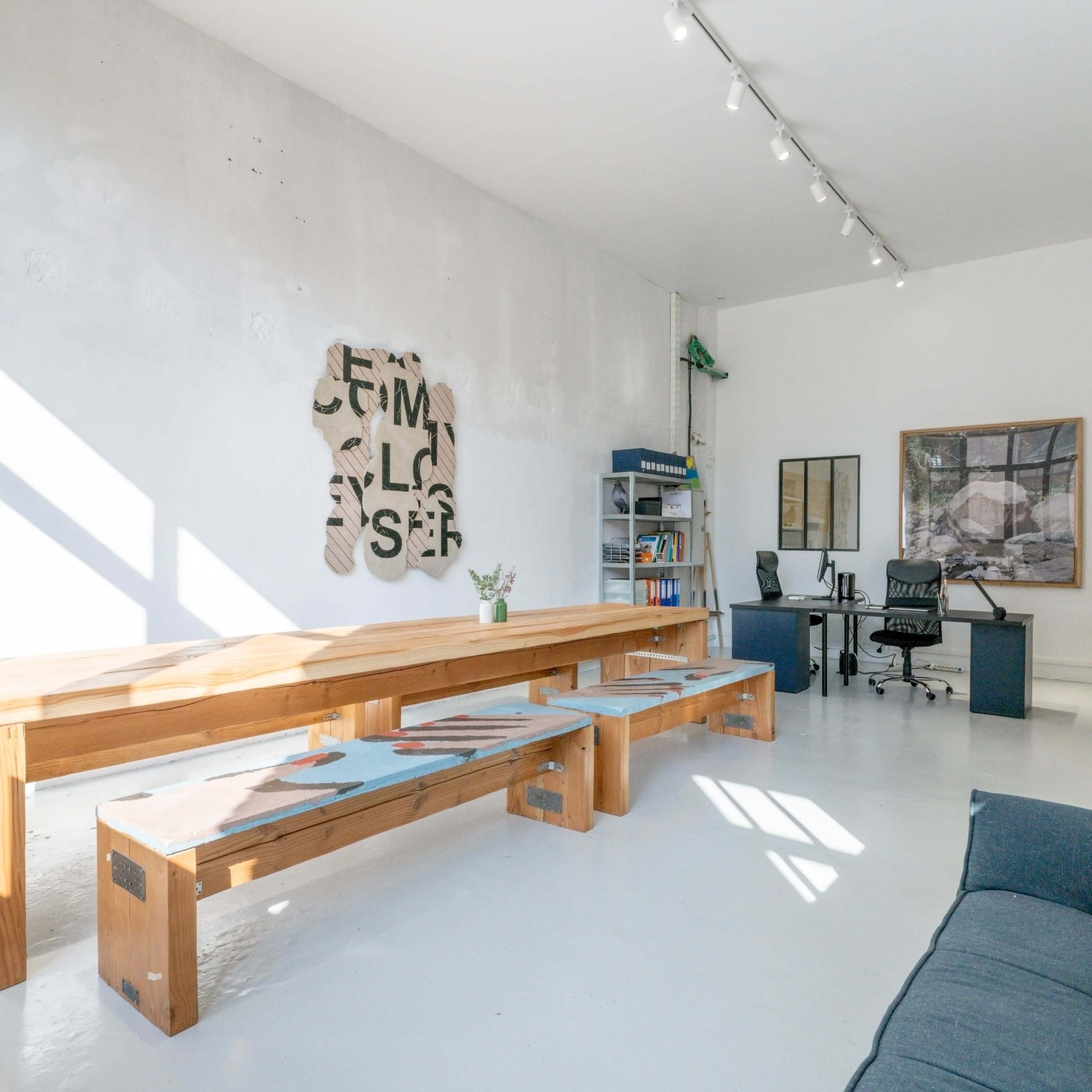 A minimalist office space featuring a large wooden table, a sofa, a desk with a chair, and a wall with abstract art.