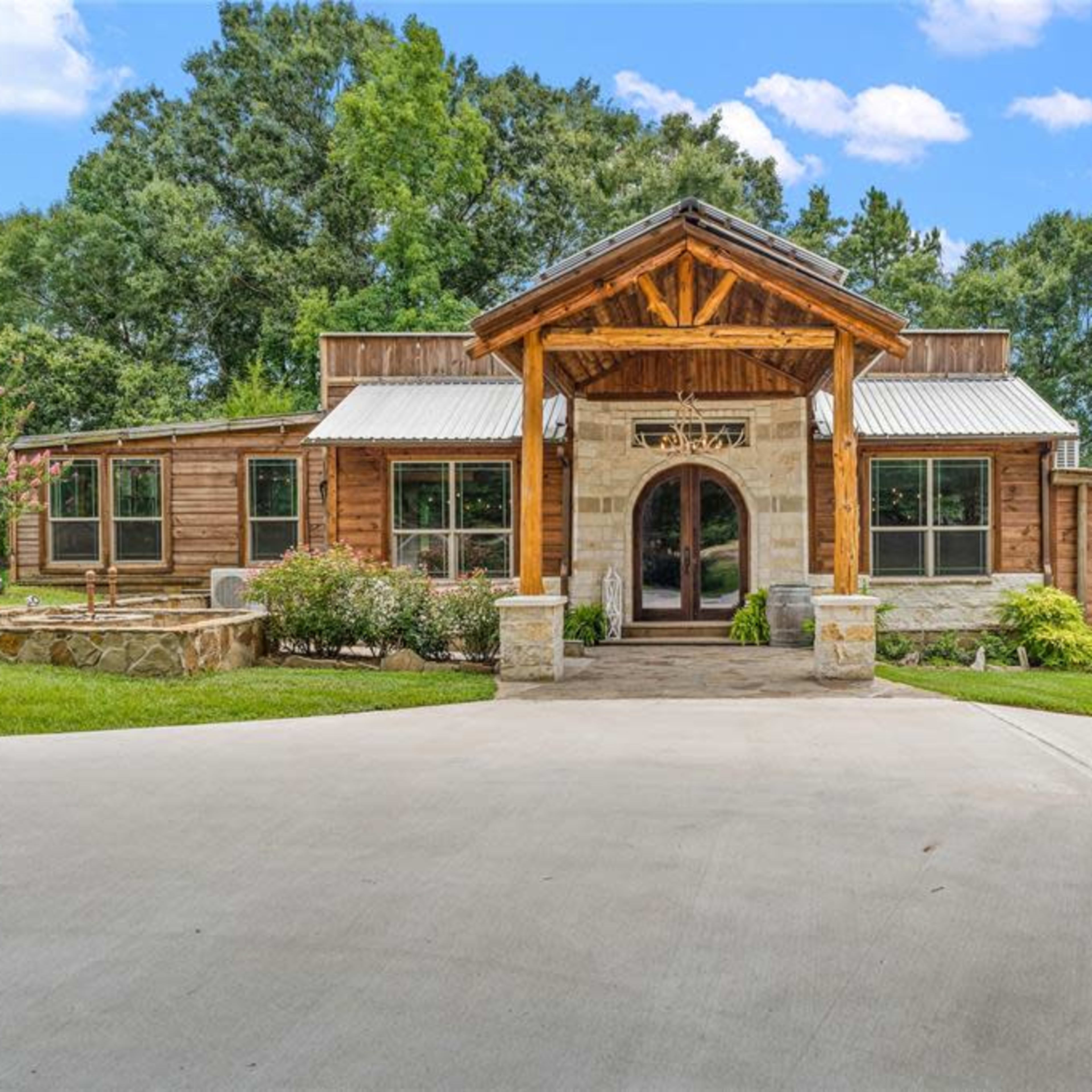 A large, wooden house with a prominent front porch and stone accents is surrounded by lush greenery.