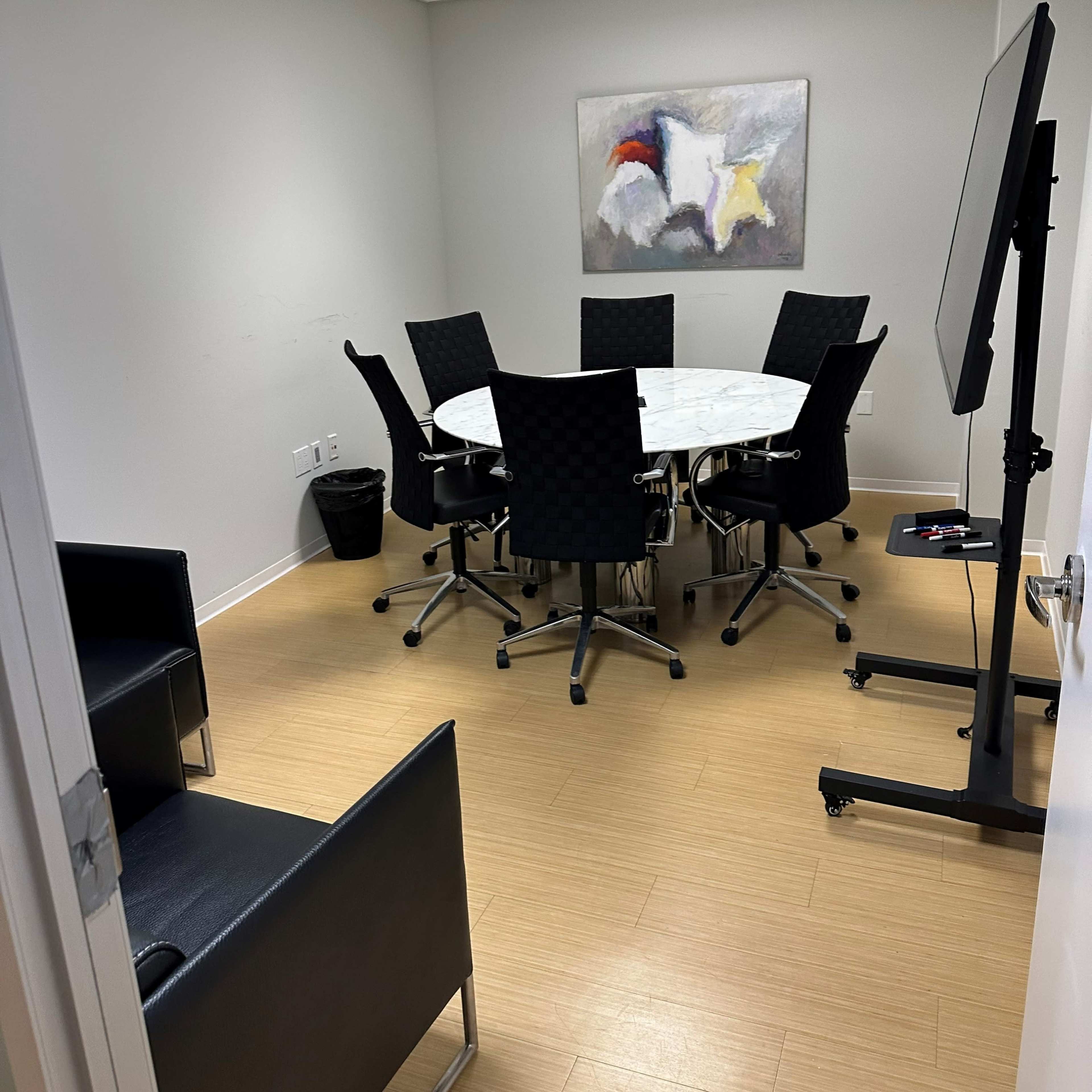 A conference room with a round table surrounded by six black chairs, an abstract painting on the wall, and a presentation screen on a stand.