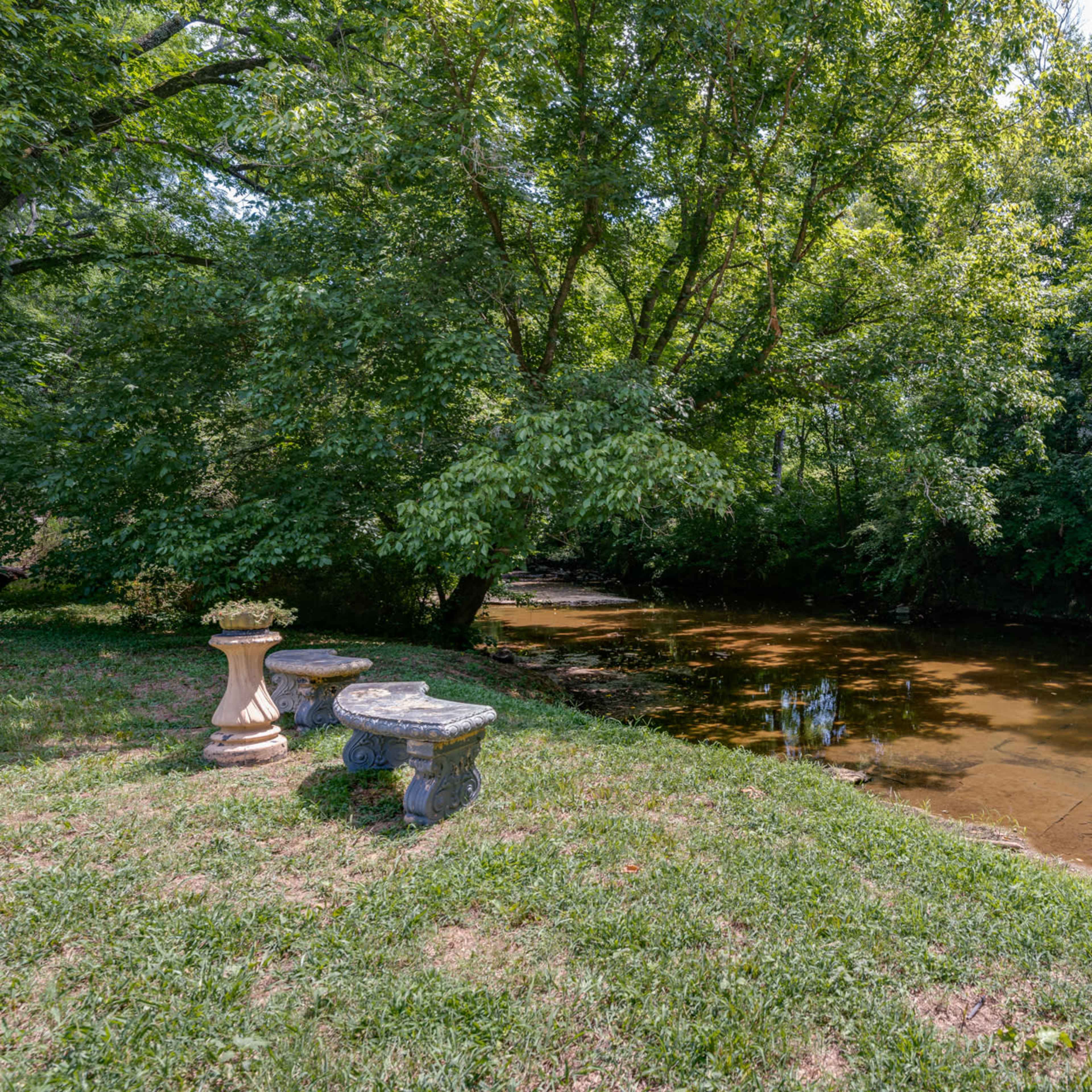 A small creek flows beside a grassy area with two stone tables surrounded by trees.