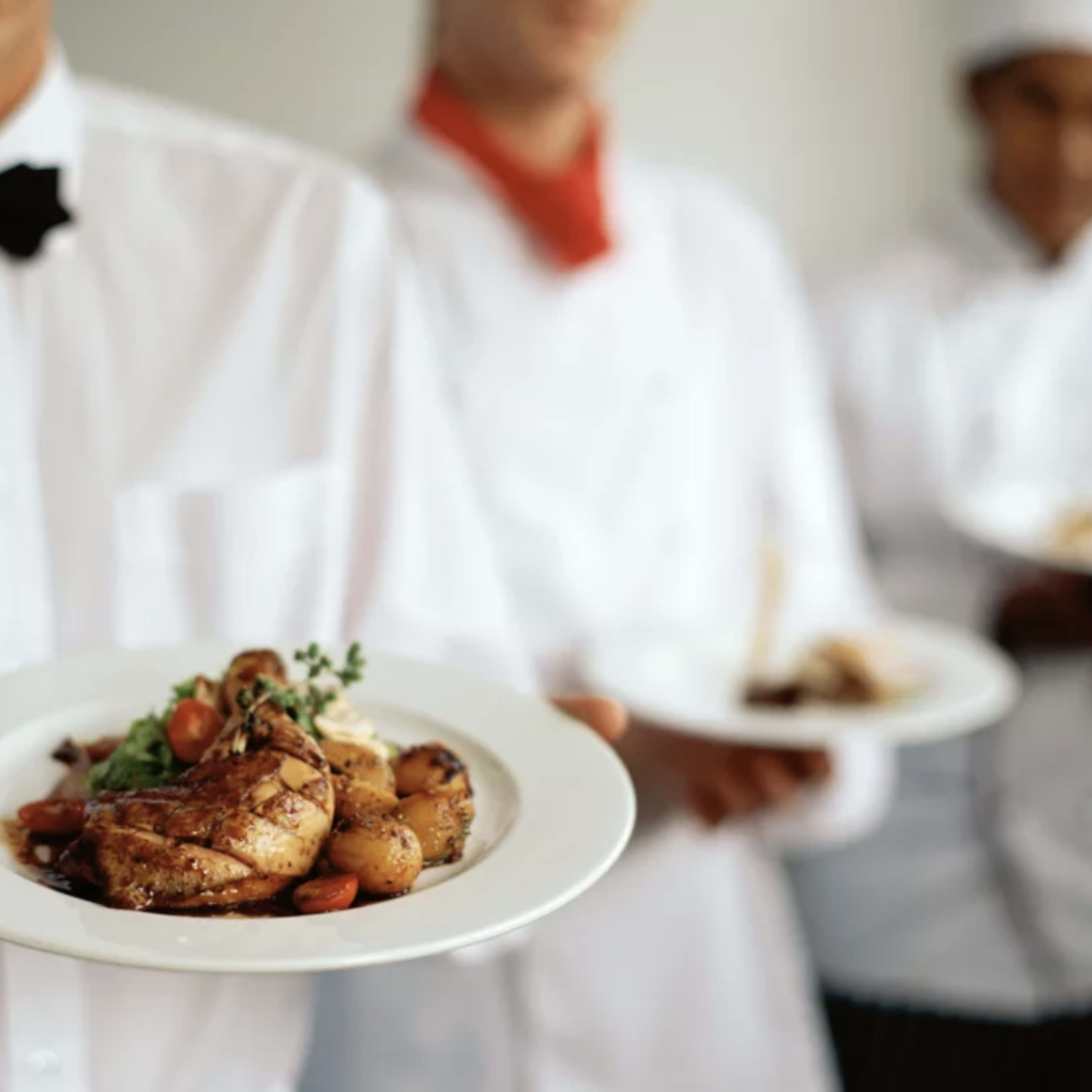 Three chefs in white uniforms hold plates of food, with one chef in the foreground presenting a dish featuring roasted chicken, potatoes, and garnishes.
