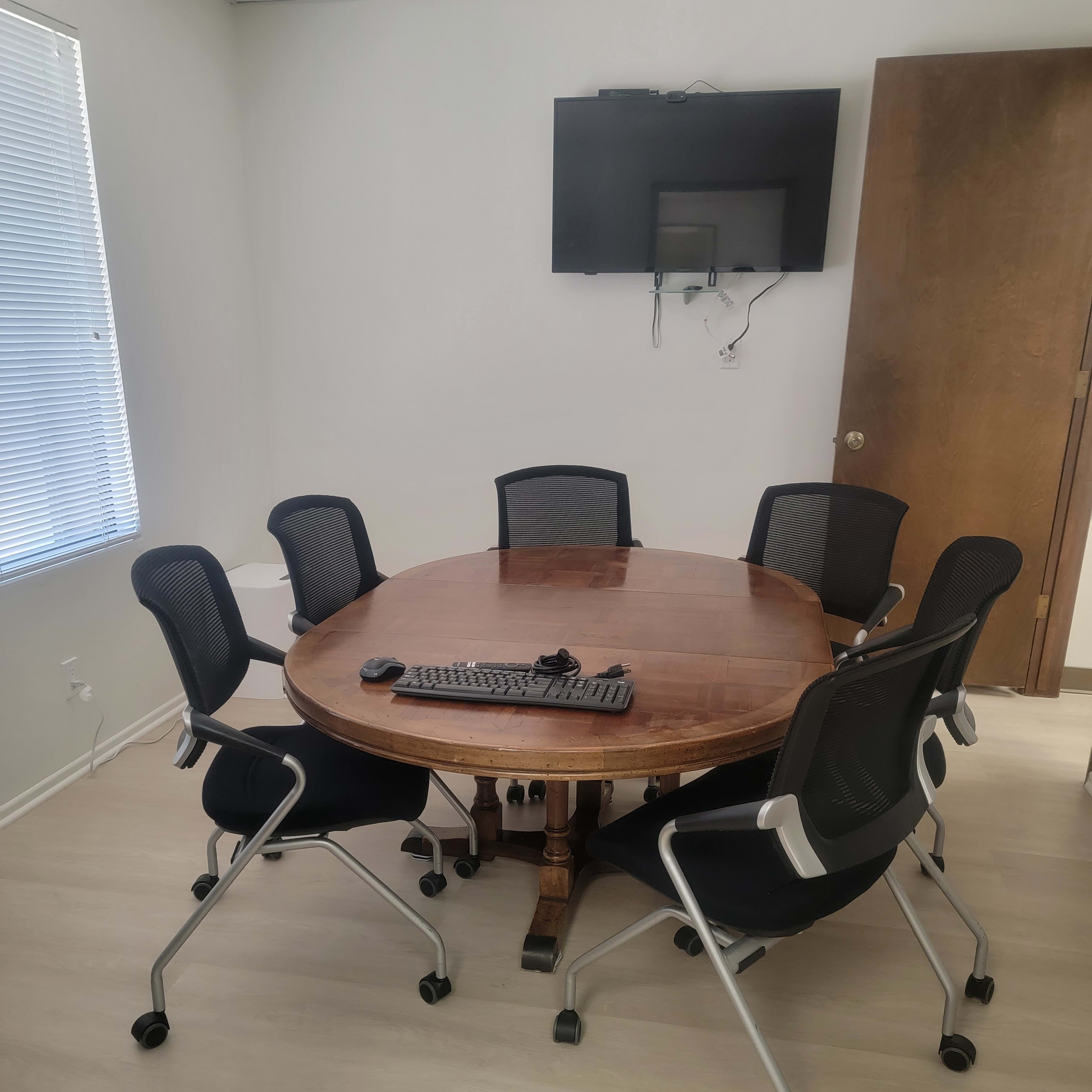 A round wooden table surrounded by six black rolling chairs is positioned in a well-lit meeting room, with a television mounted on the wall.