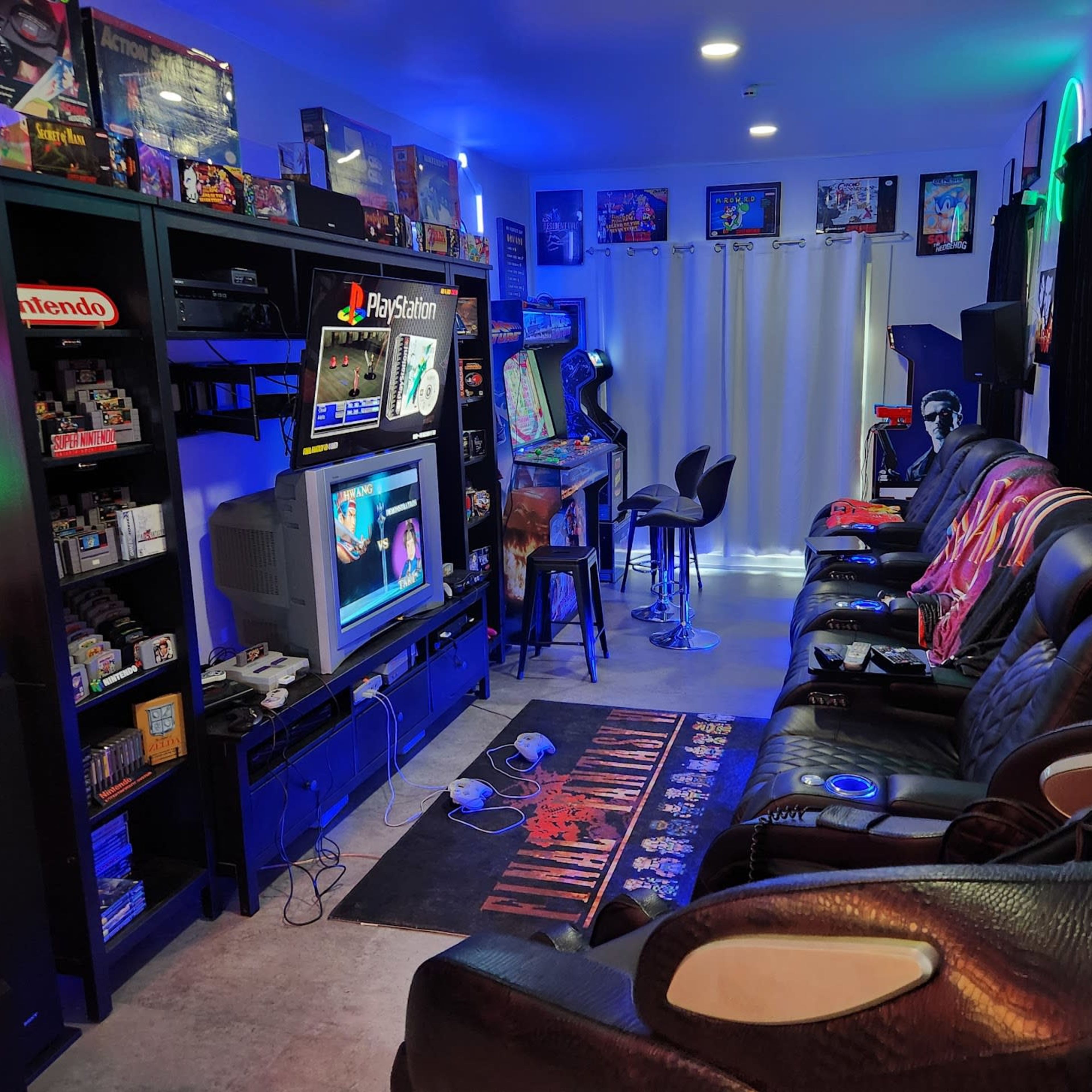 A gaming room with black leather seating, a large shelf filled with video game consoles and memorabilia, and a television displaying a game, all illuminated by colorful lights.