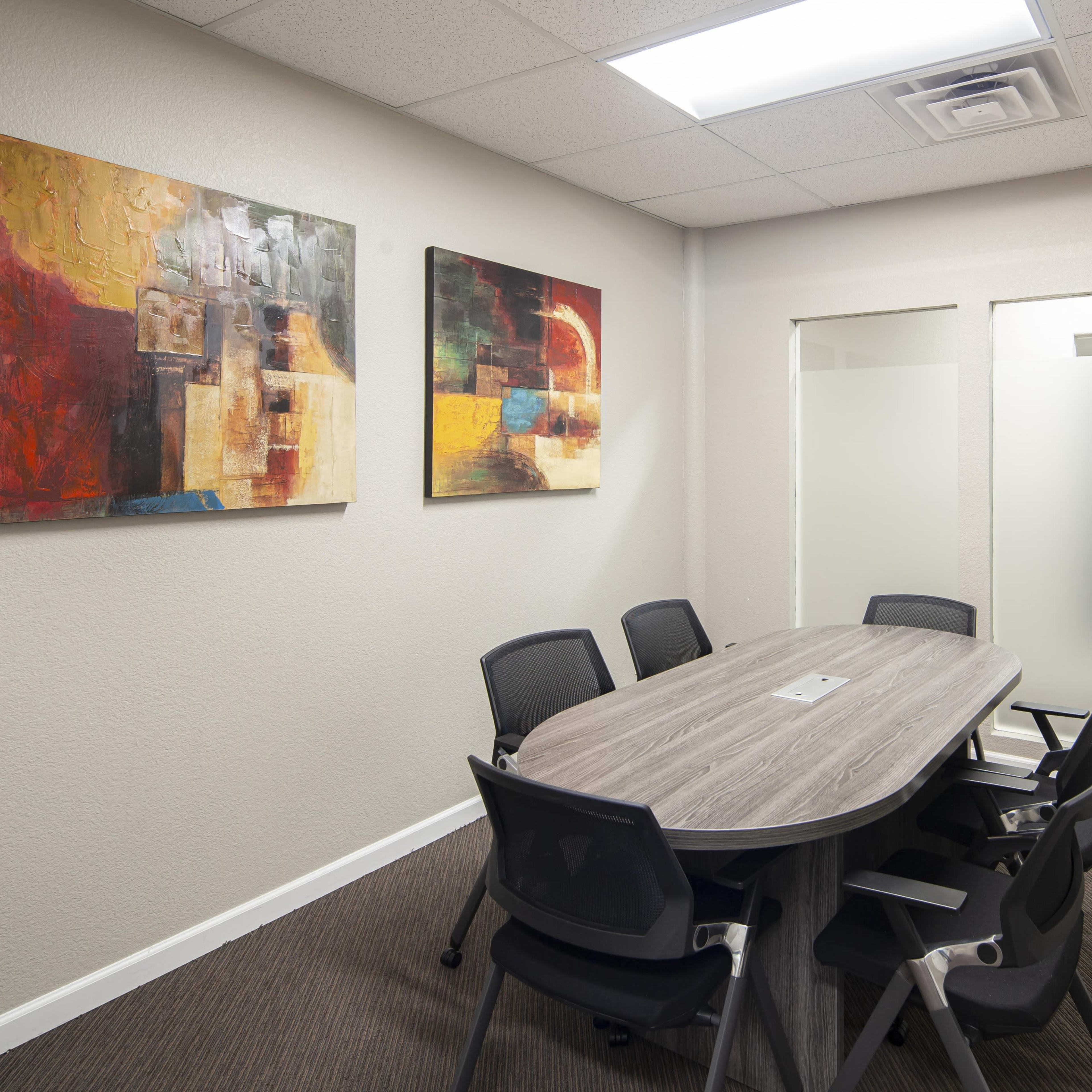 A conference room features a large oval table surrounded by black chairs, with abstract paintings on the walls and glass-paneled doors.