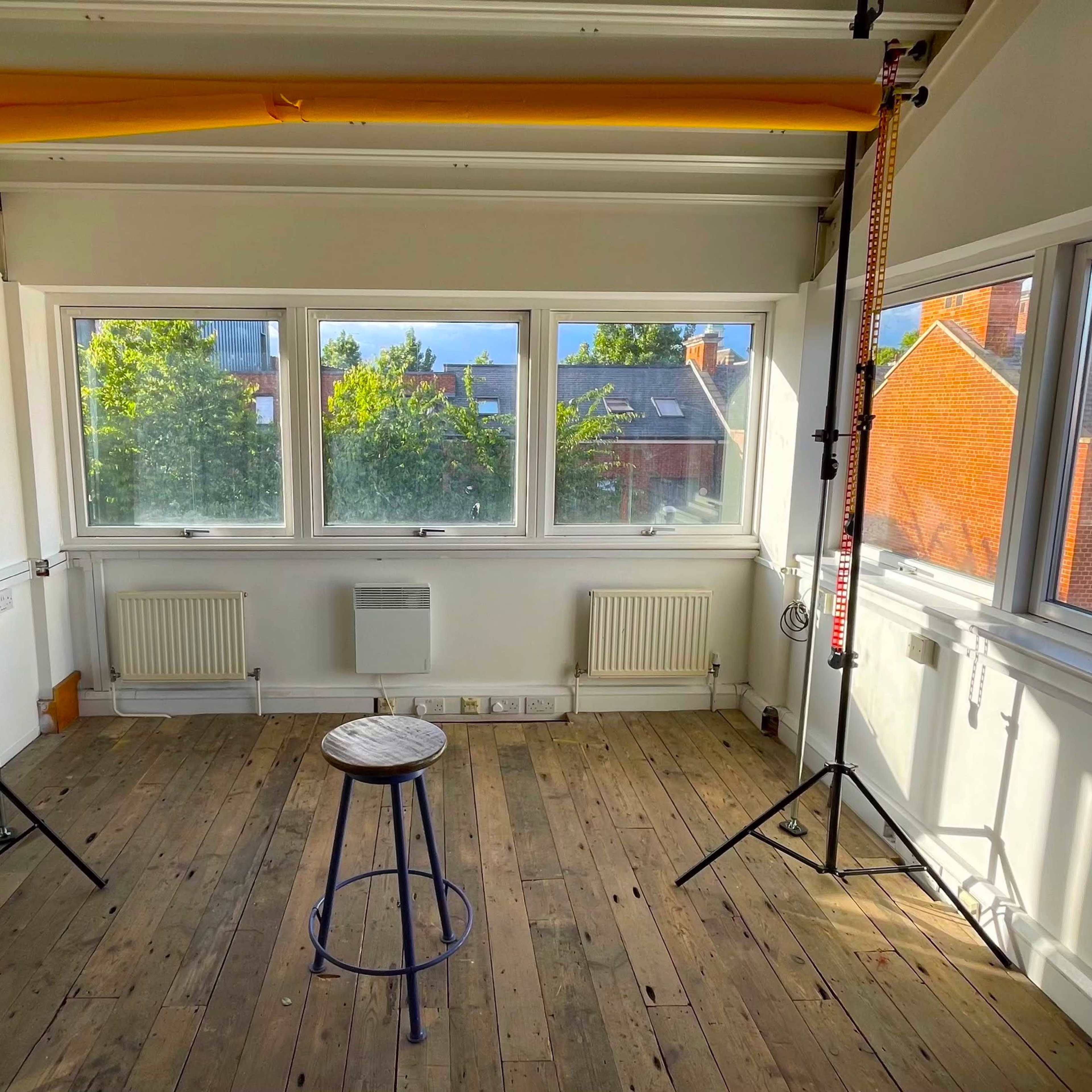 A brightly lit room with wooden flooring, featuring a stool centered under two lighting stands and large windows.