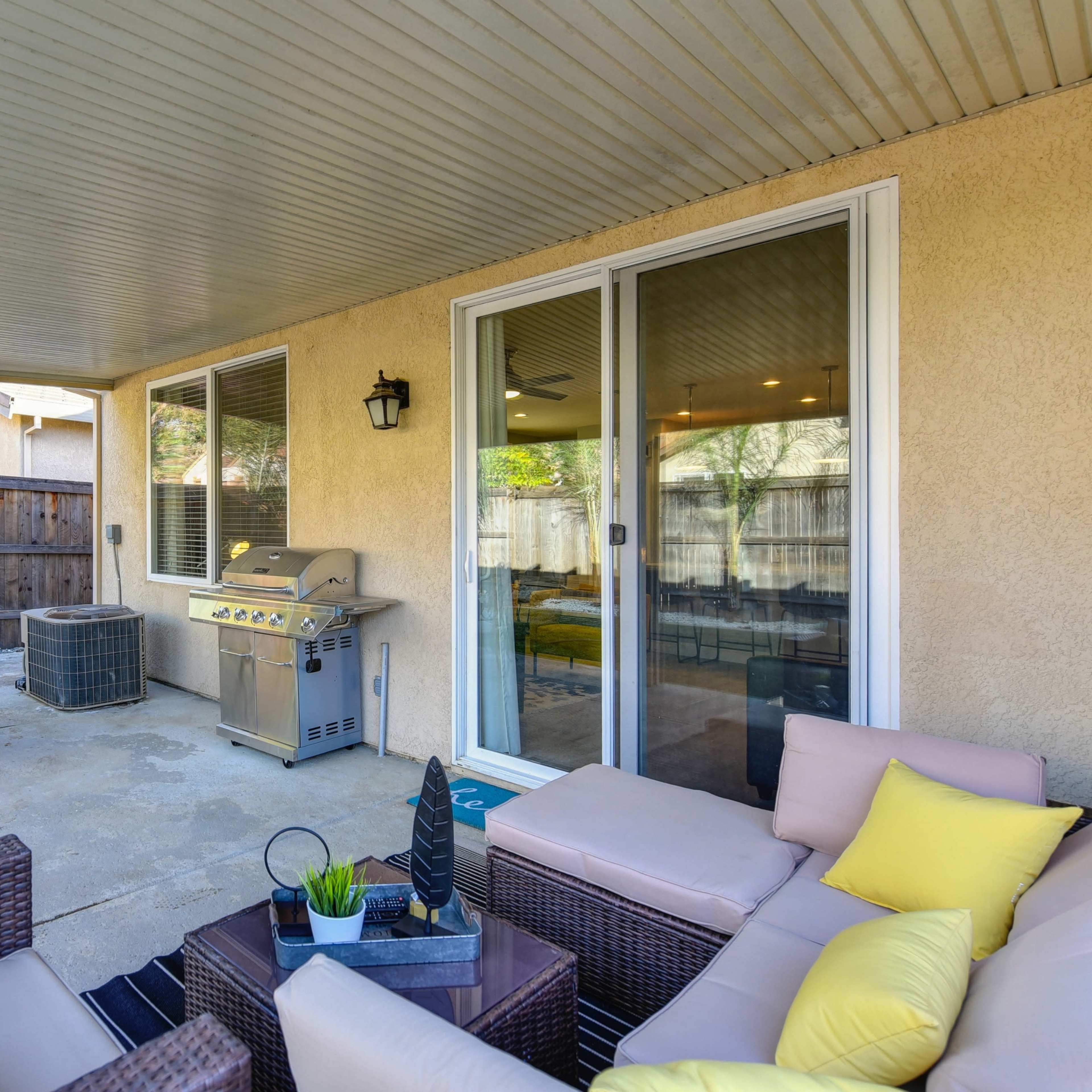A covered patio features a seating area with a table, grill, and an air conditioning unit surrounded by a wooden fence.