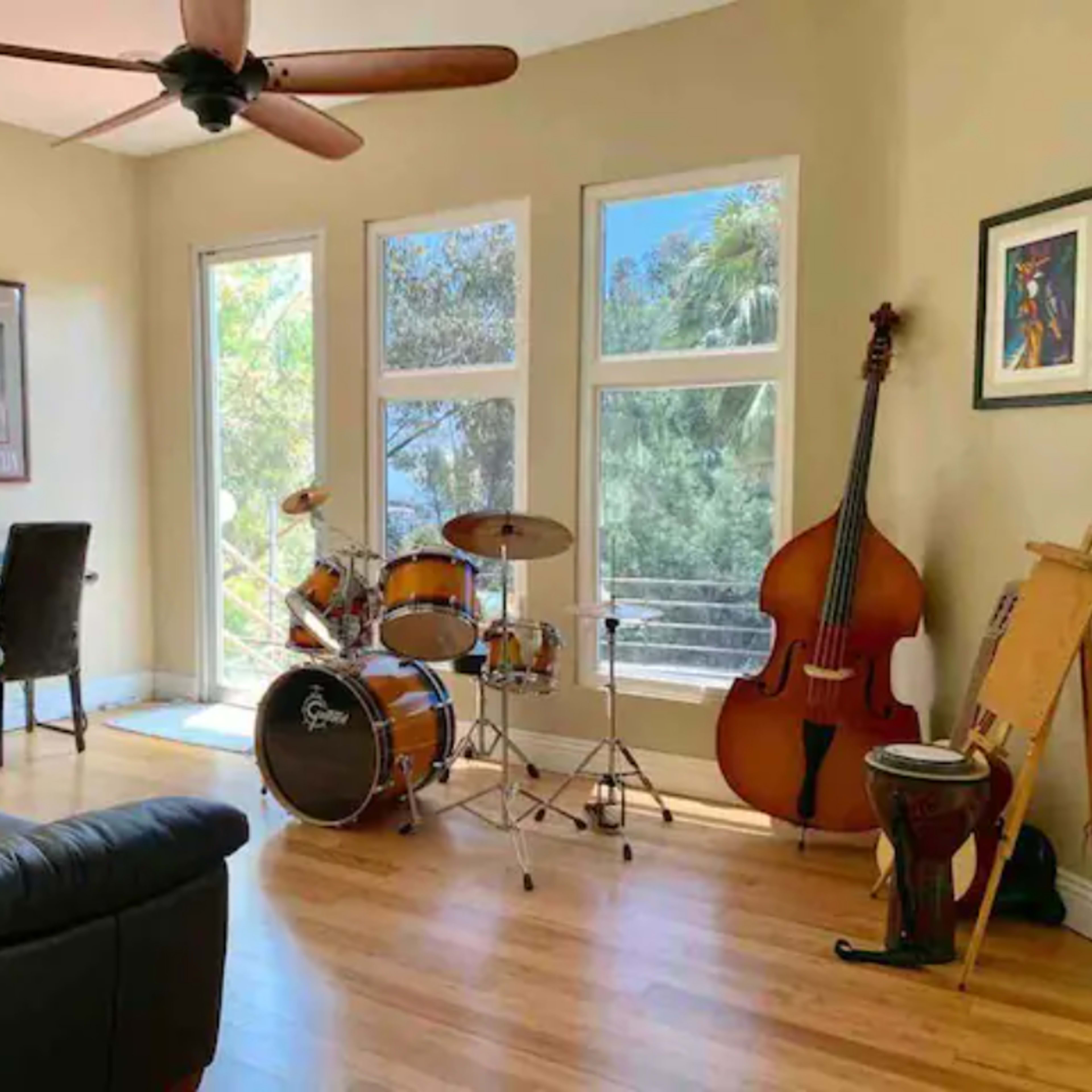 A musical room featuring a black leather couch, a drum set, a double bass, and an easel, with large windows providing a view of greenery outside.