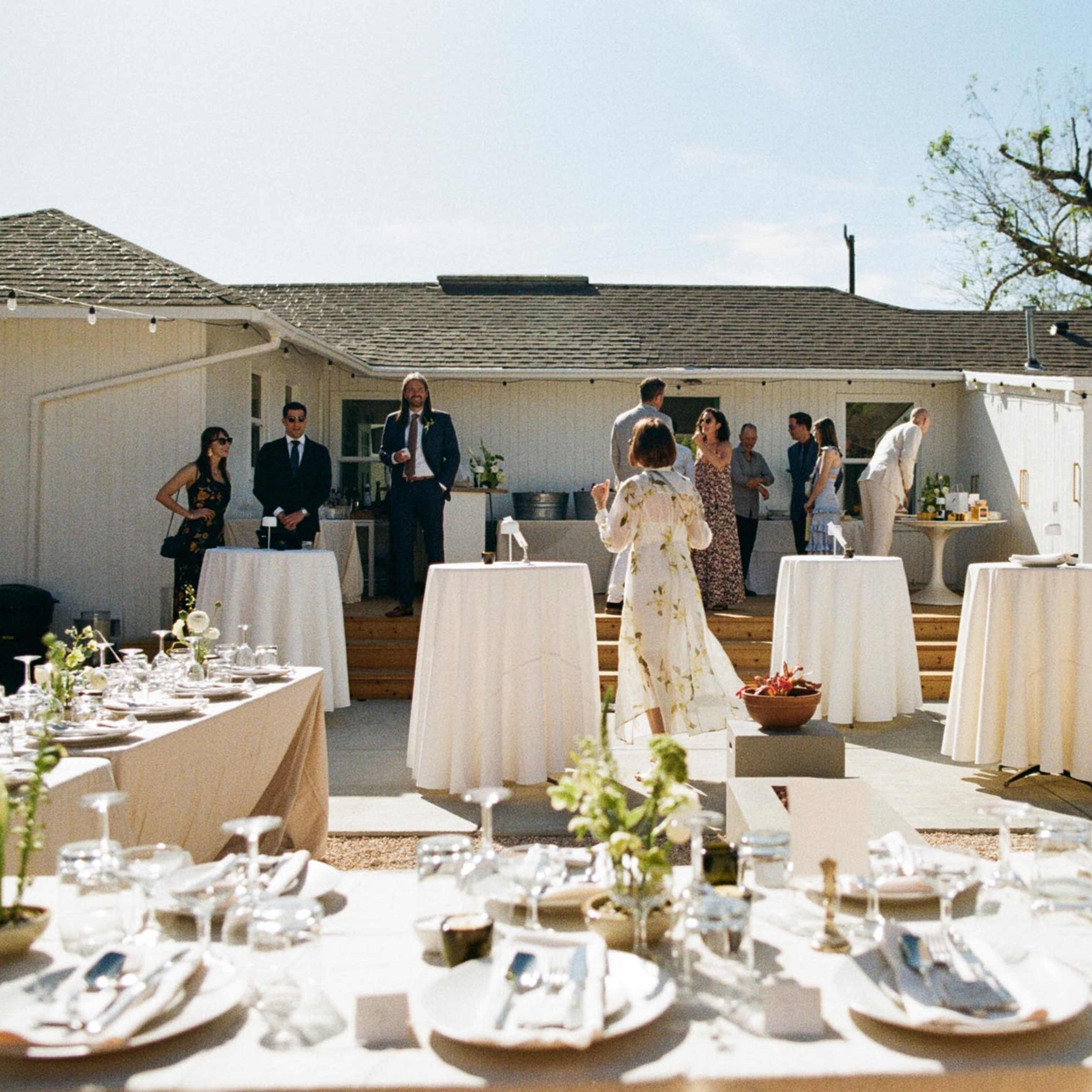 An outdoor gathering is set with several dining tables, surrounded by guests and a house in the background.