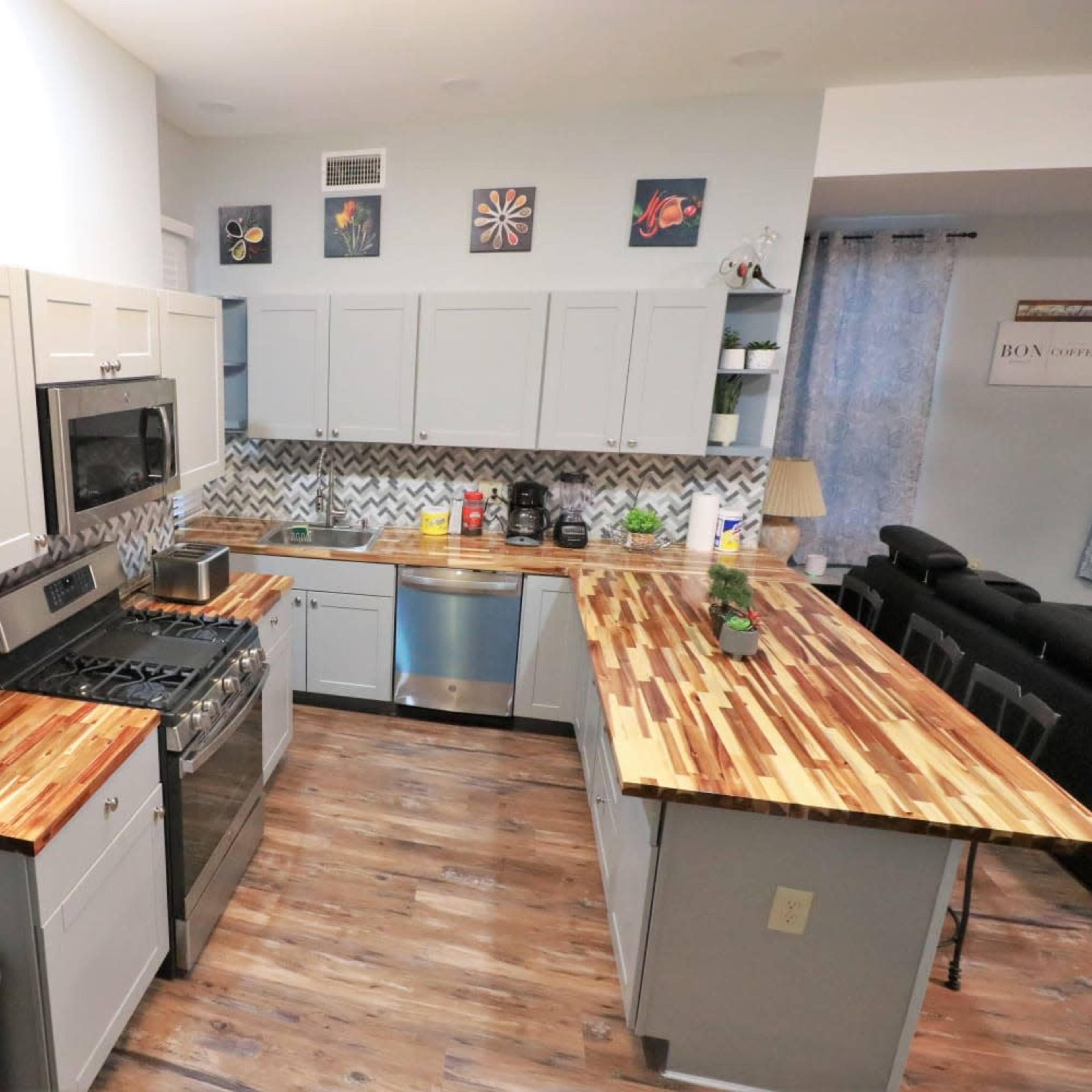 A modern kitchen features light gray cabinets, stainless steel appliances, and a wooden countertop with a chevron-patterned backsplash.