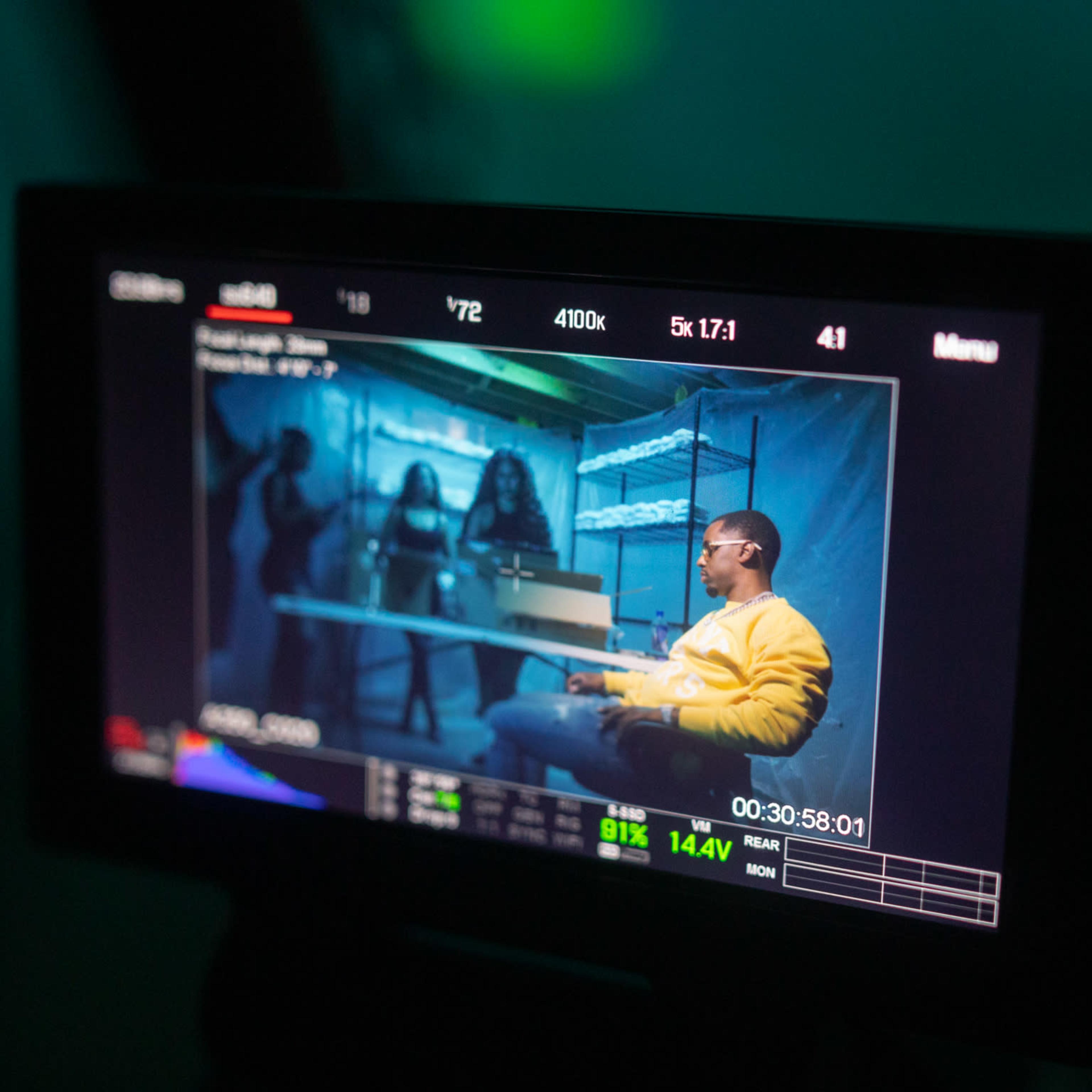 A monitor displaying a scene from a film set featuring a person sitting at a table while three others stand in the background.