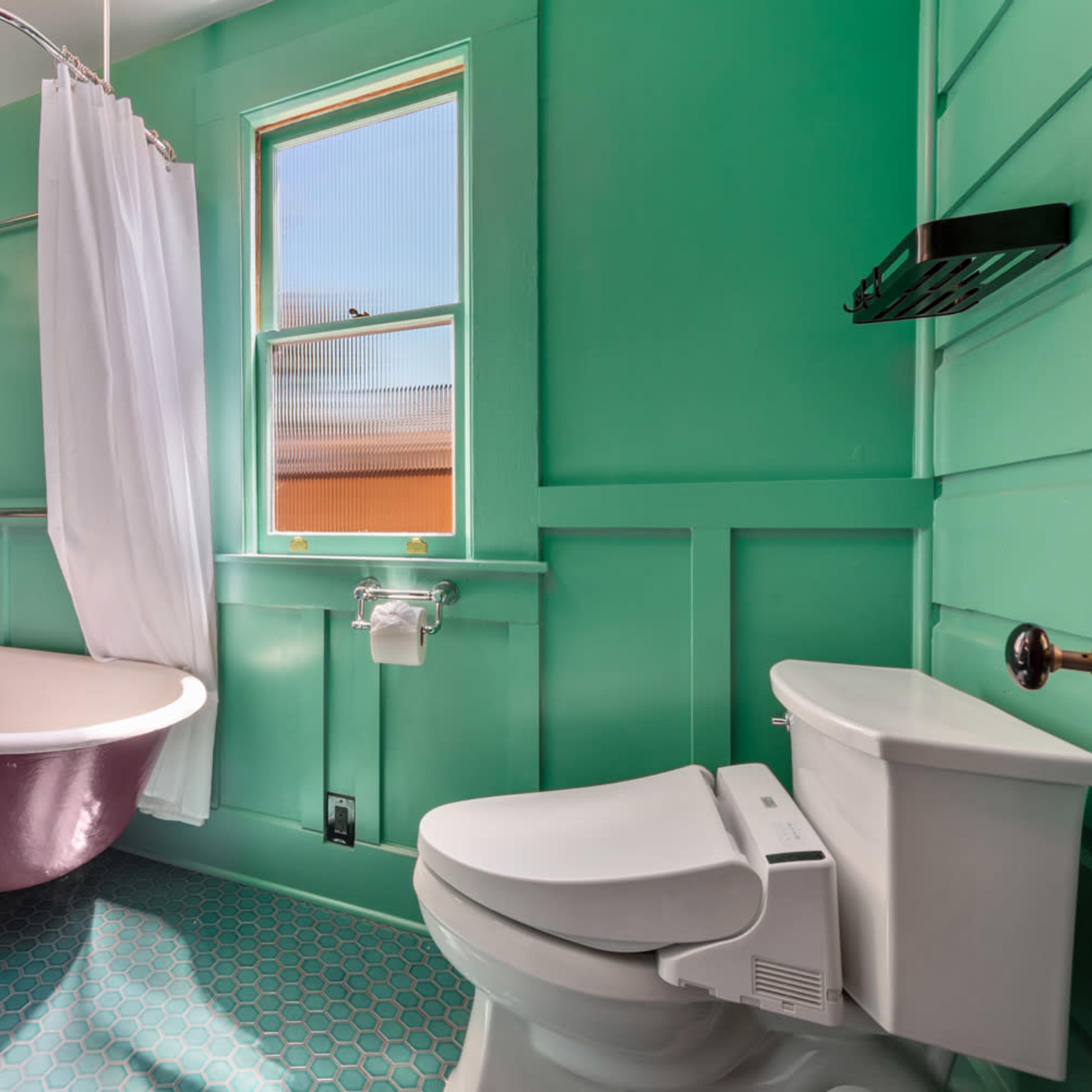 A bathroom with green walls, a freestanding clawfoot bathtub, a shower curtain, and a toilet, along with a window allowing natural light.