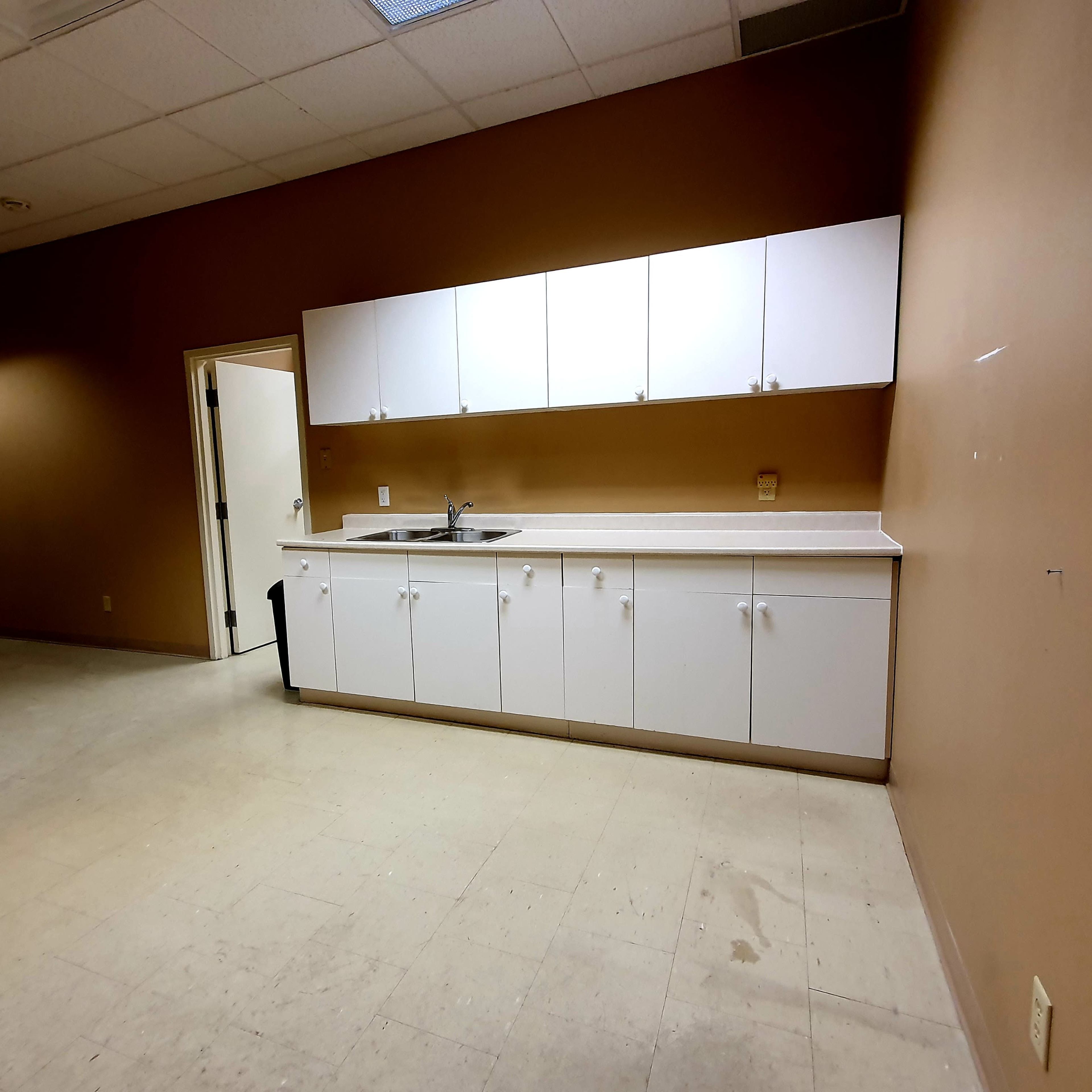 A small kitchen area with white cabinets and a sink against a brown wall in a mostly empty room.