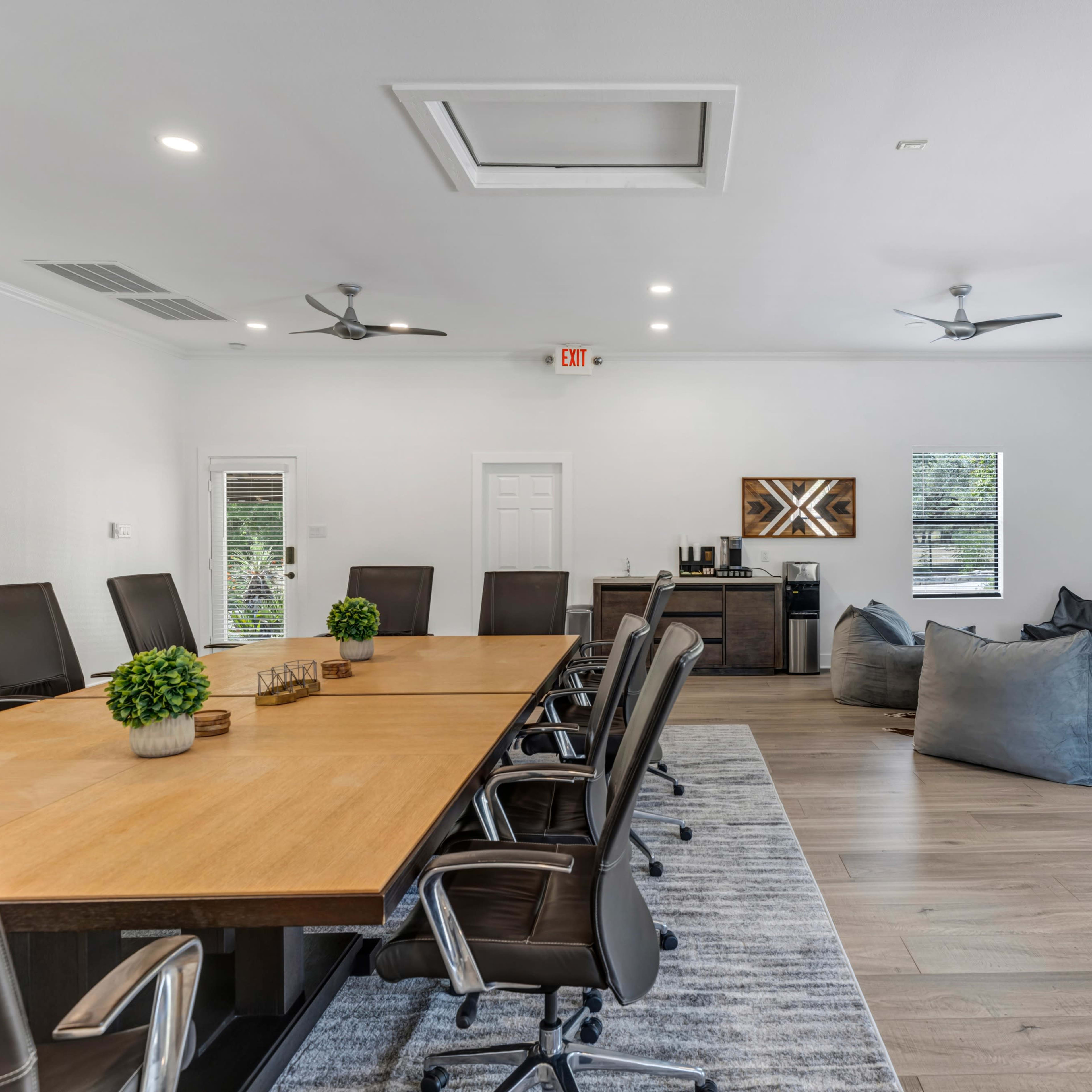 A spacious conference room features a long wooden table surrounded by black office chairs, with bean bag seating and a coffee area against the walls.