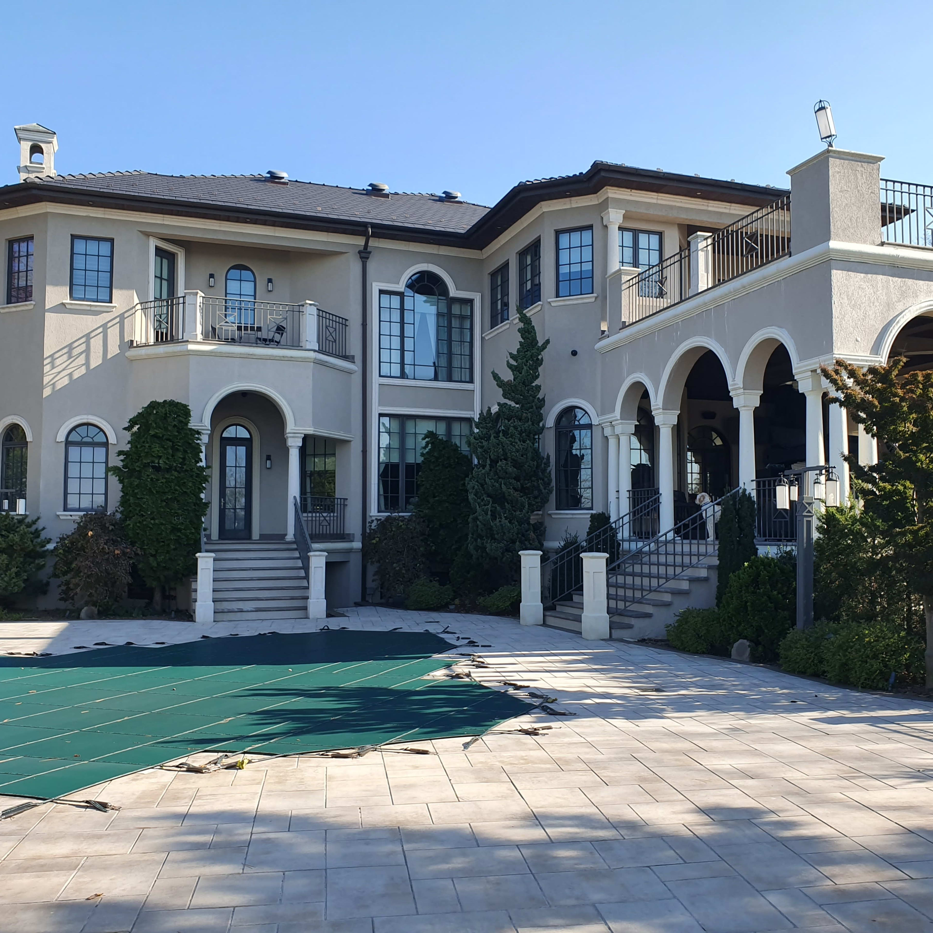 A large, two-story house with a mix of arched and rectangular windows, a balcony, and a stone walkway, set against a clear sky.
