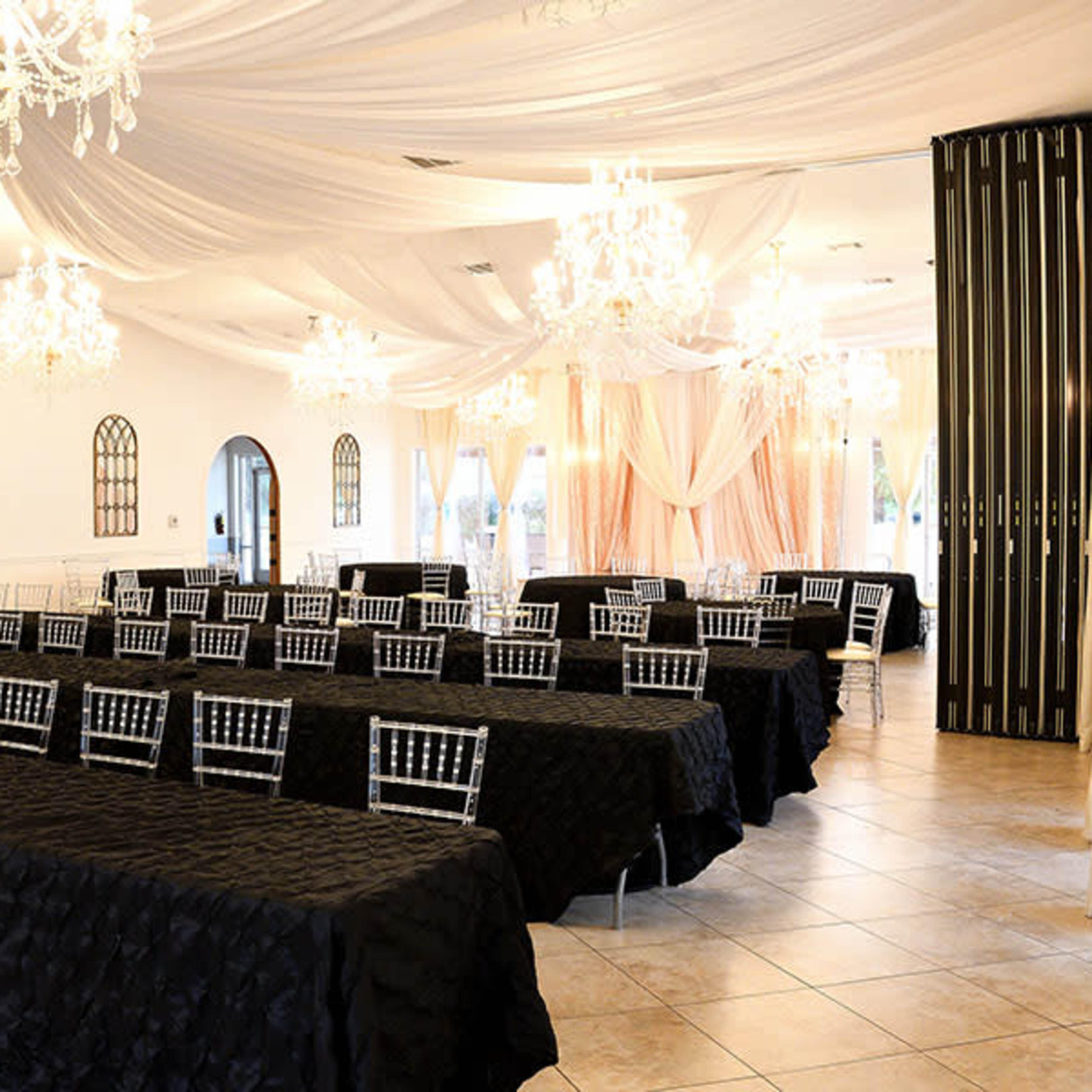 A banquet hall arranged with rows of tables covered in black tablecloths and adorned with silver Chiavari chairs under draped fabric and chandeliers.