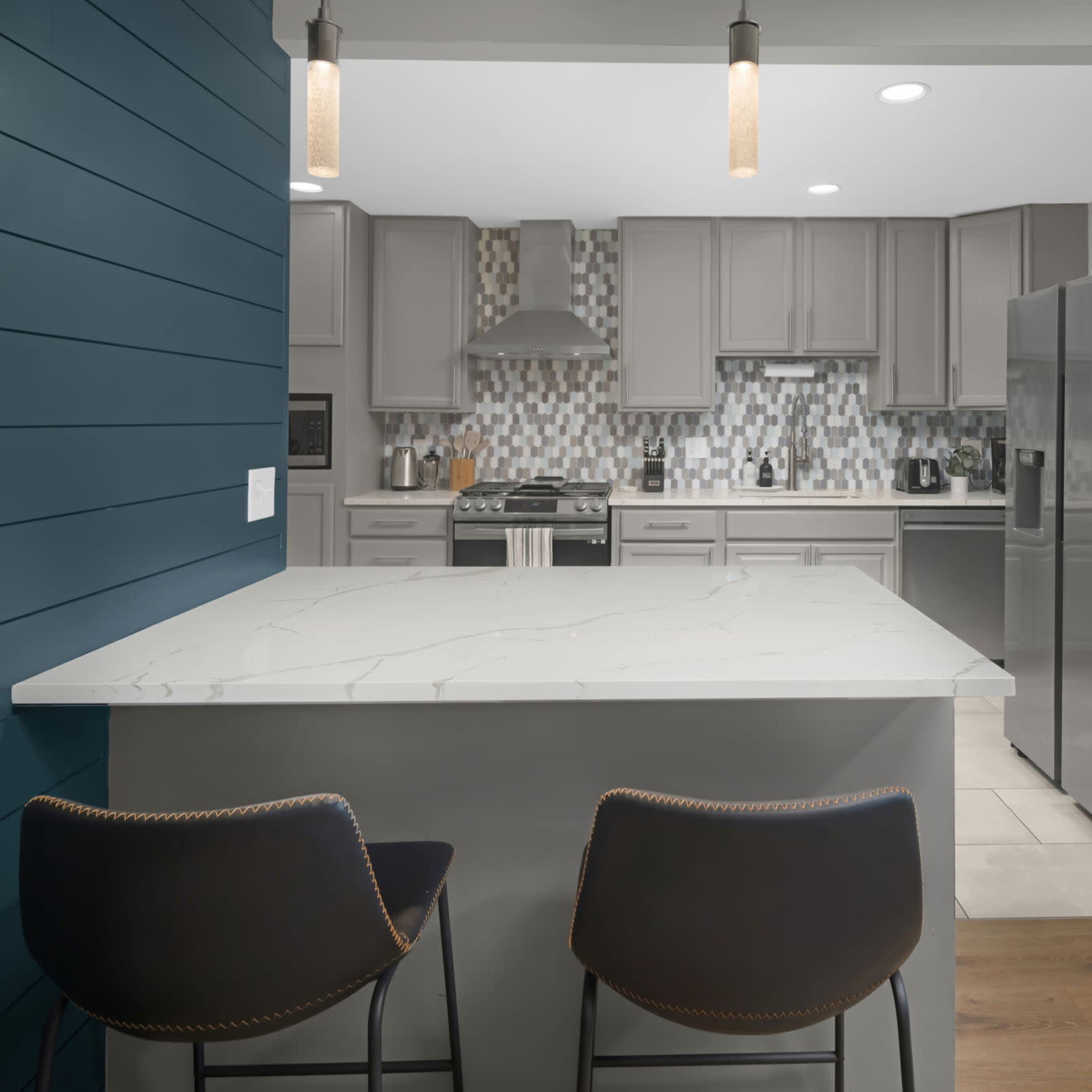 A modern kitchen with gray cabinets, a mosaic tile backsplash, and bar stools at a marble countertop.