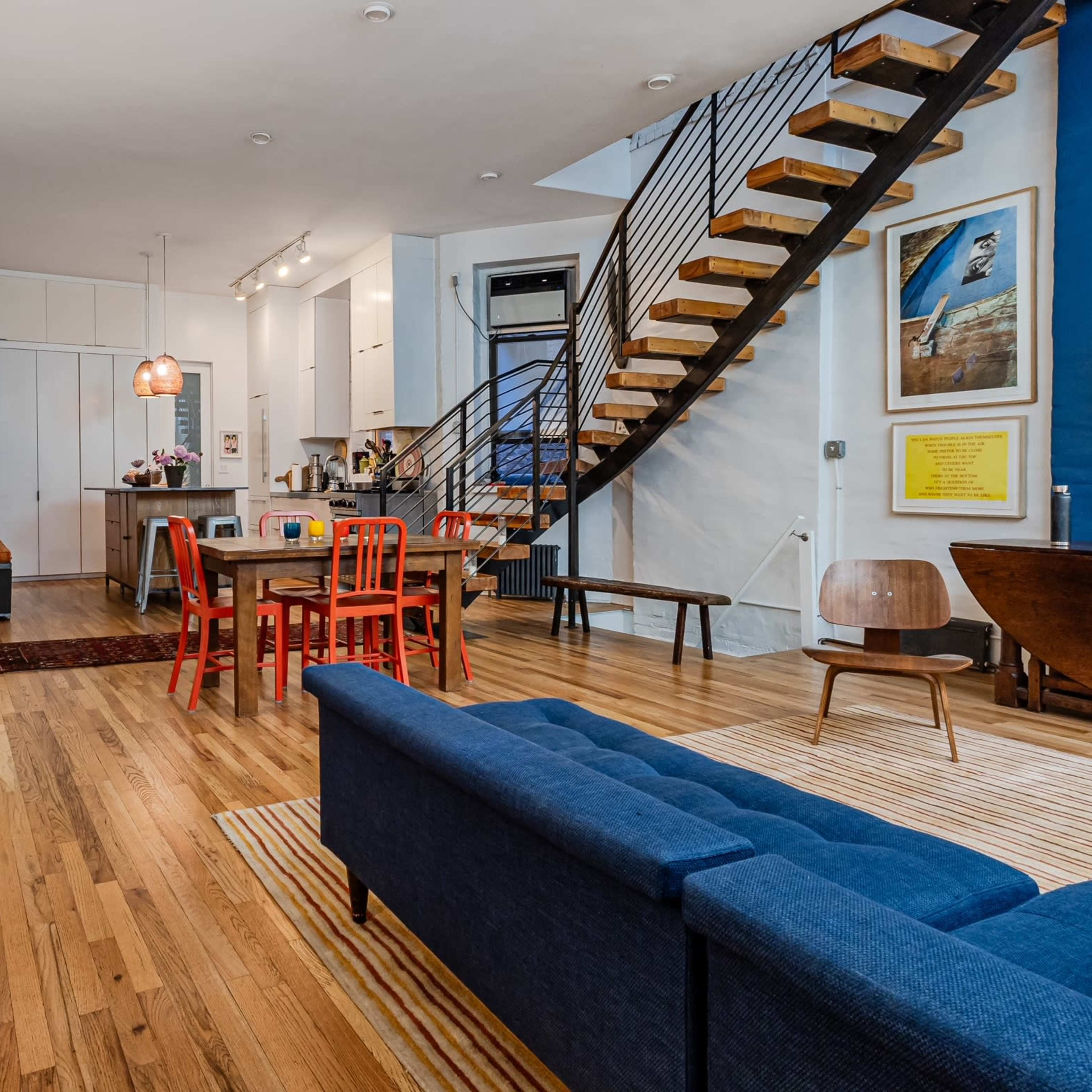 A modern living space with a blue sofa, a wooden dining table surrounded by red chairs, a staircase leading to an upper level, and various artworks on the walls.