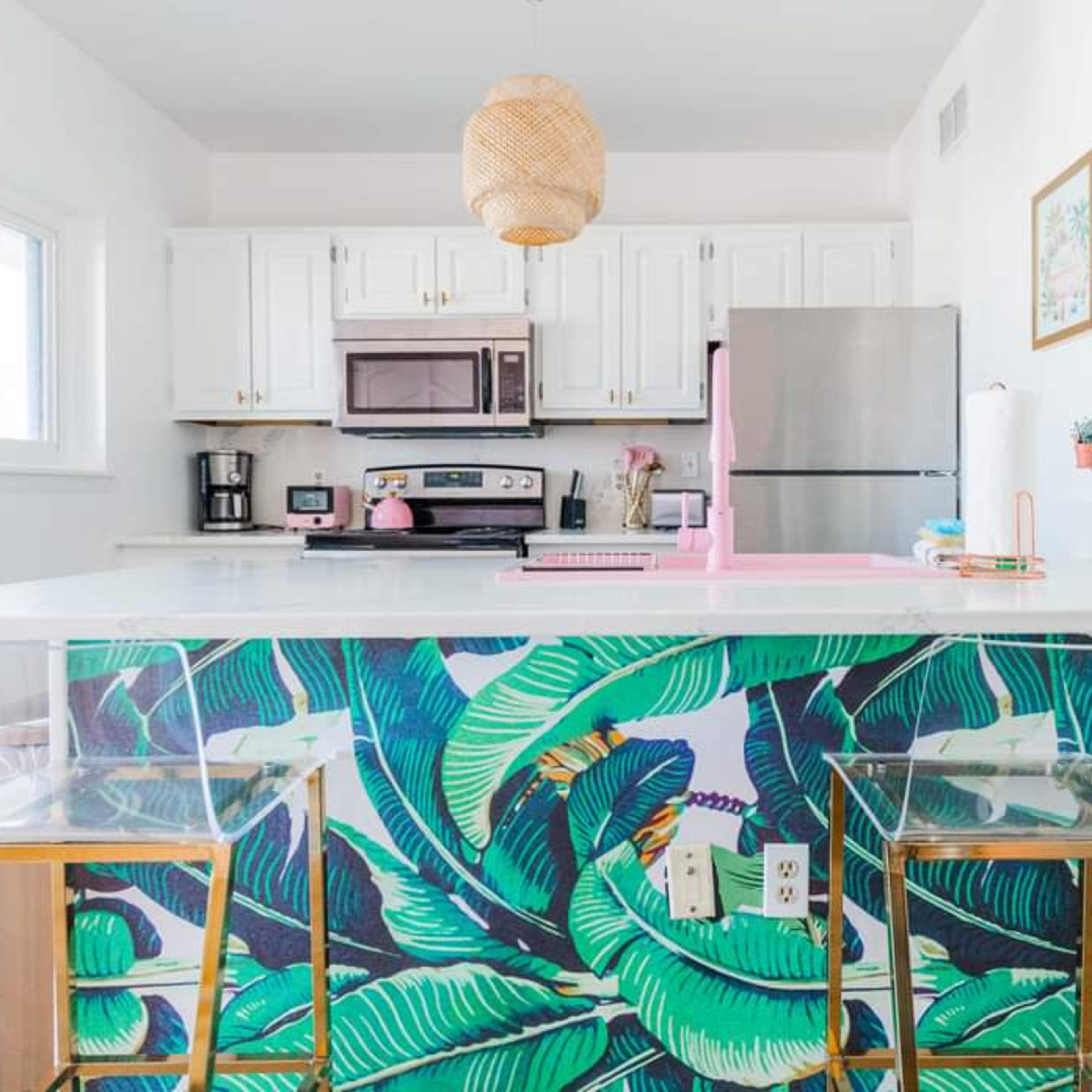 A modern kitchen features white cabinetry, stainless steel appliances, and a bar area with a tropical-themed backsplash and two clear chairs.