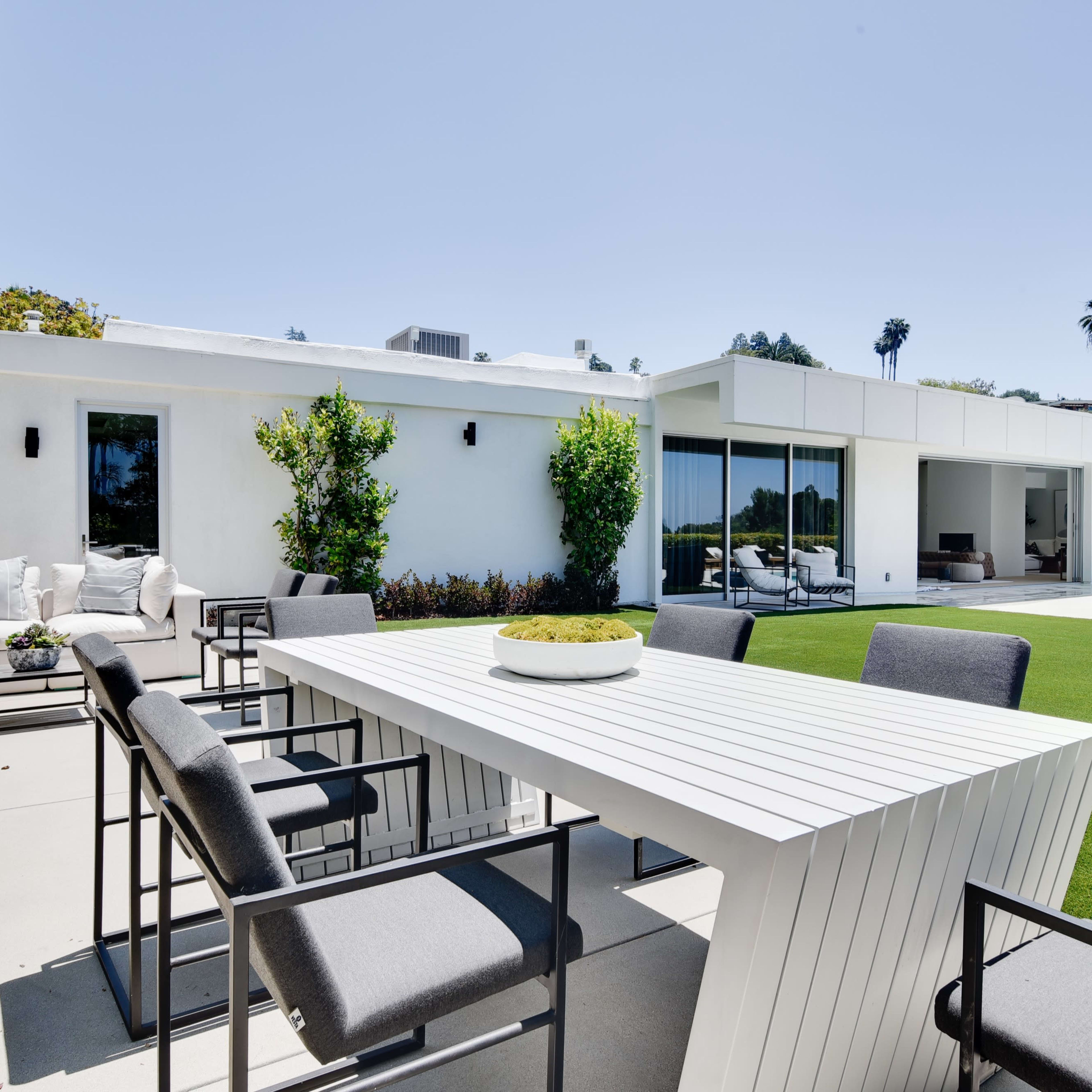 A modern outdoor dining area features a white table surrounded by gray chairs, adjacent to a swimming pool and a neatly maintained lawn.