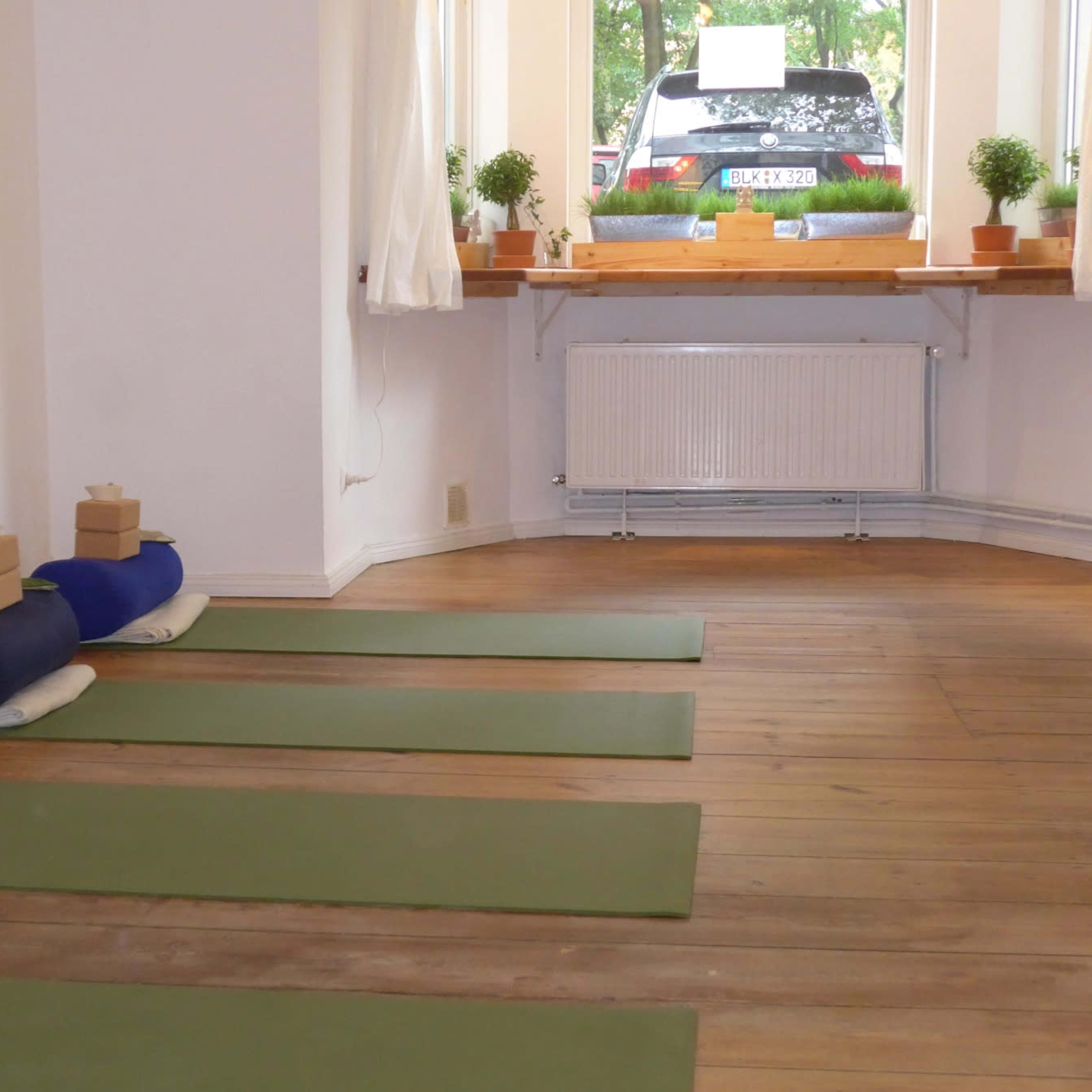 A yoga studio with green mats arranged on a wooden floor, blocks placed on top of rolled mats, and a car visible through a window.
