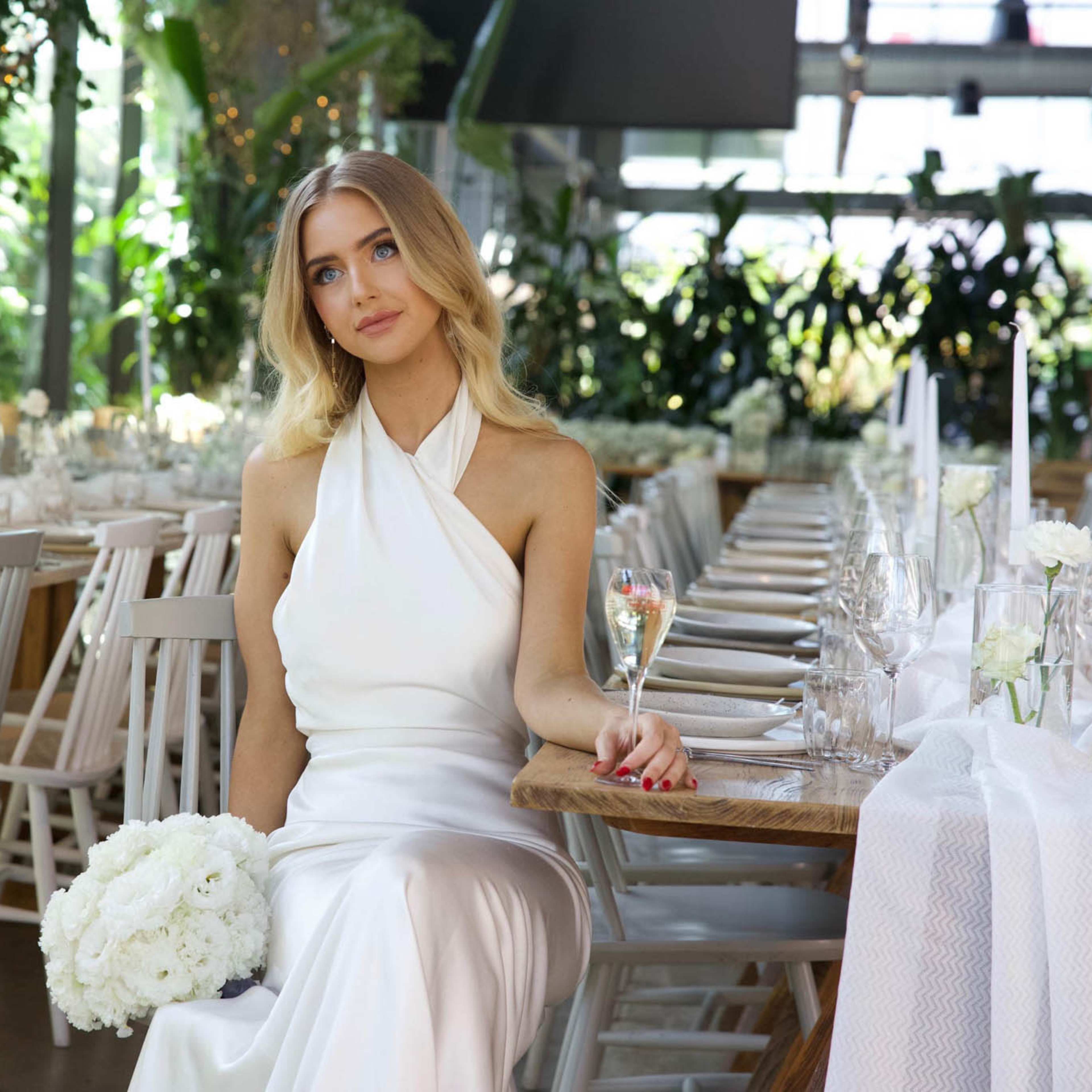 A woman in a white dress sits at a table set for an elegant event, holding a glass and a bouquet of white flowers.