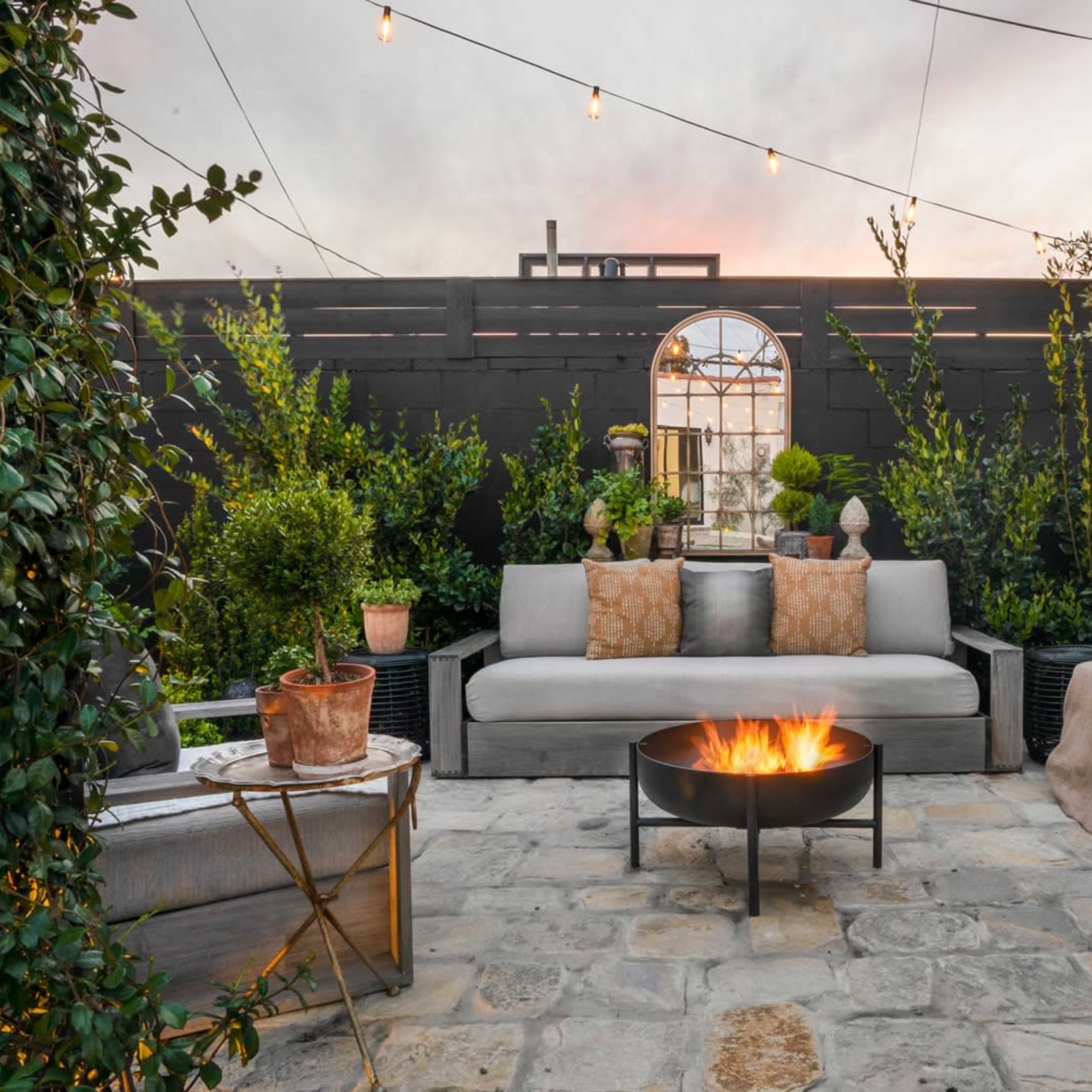 A cozy outdoor seating area with a fire pit, surrounded by plants and decorative elements under a string of lights.