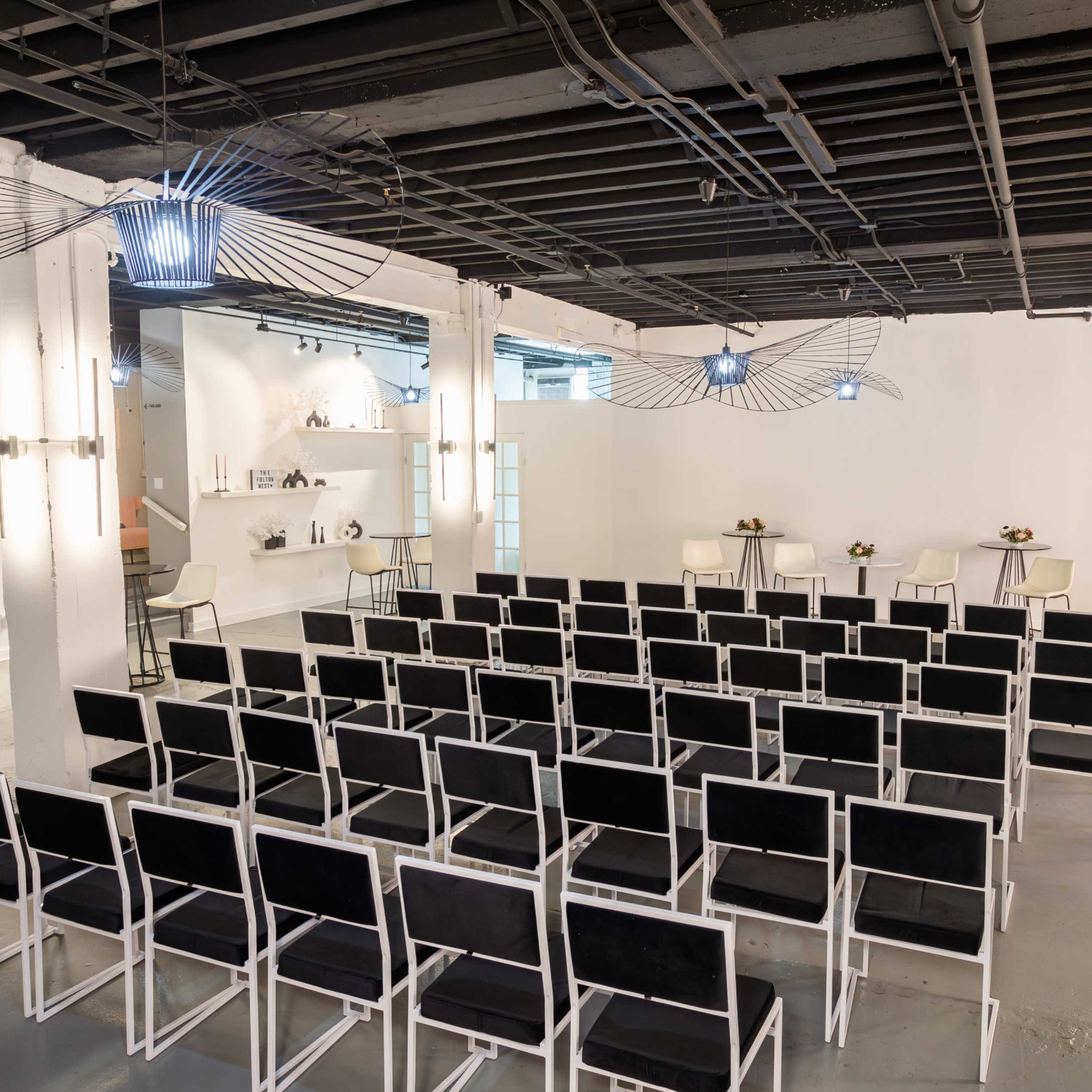 A spacious room features rows of black chairs arranged facing a minimalistic counter with two stools, illuminated by modern lighting fixtures.