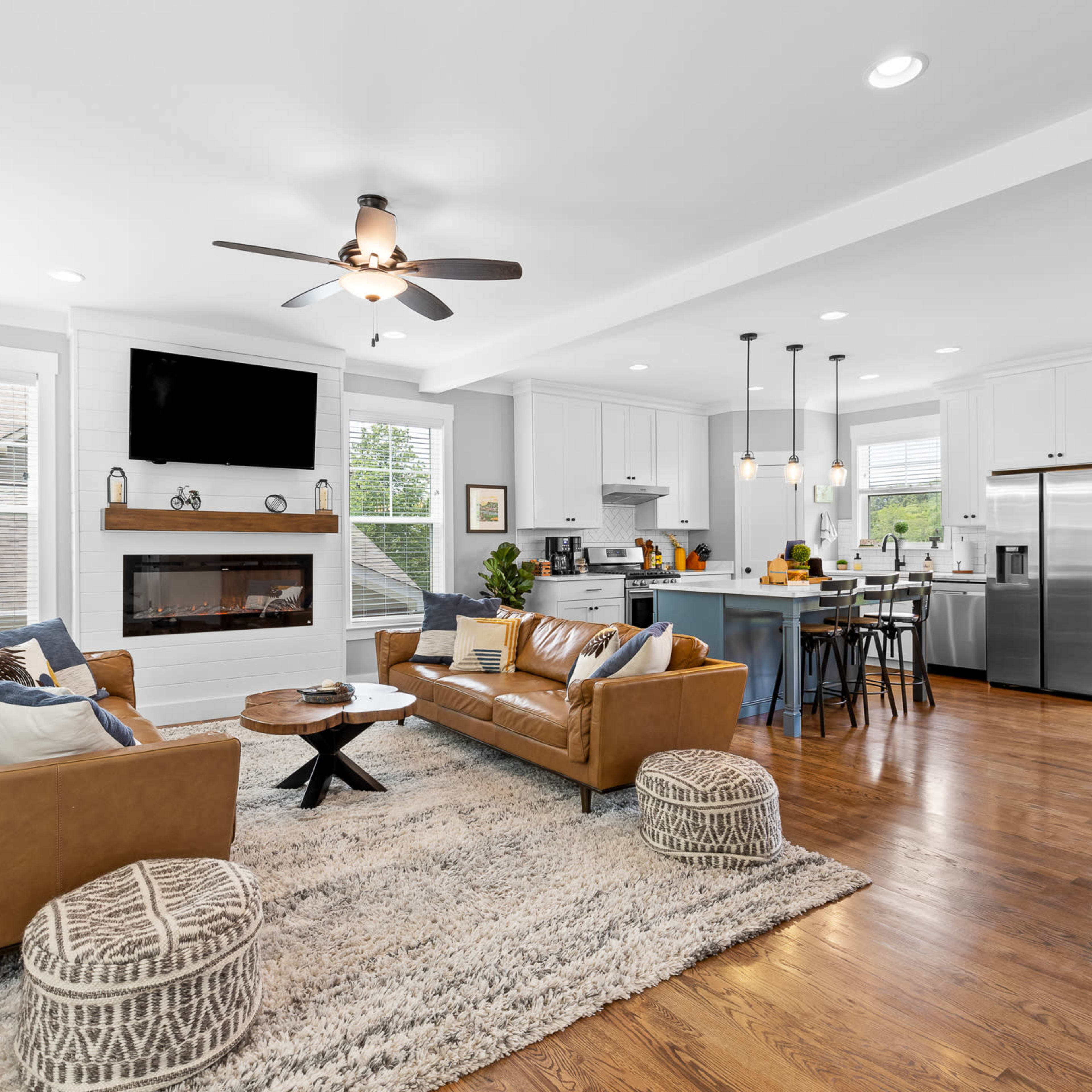 A modern living room and kitchen area, featuring a brown leather sofa, a large rug, and a kitchen island with bar stools.