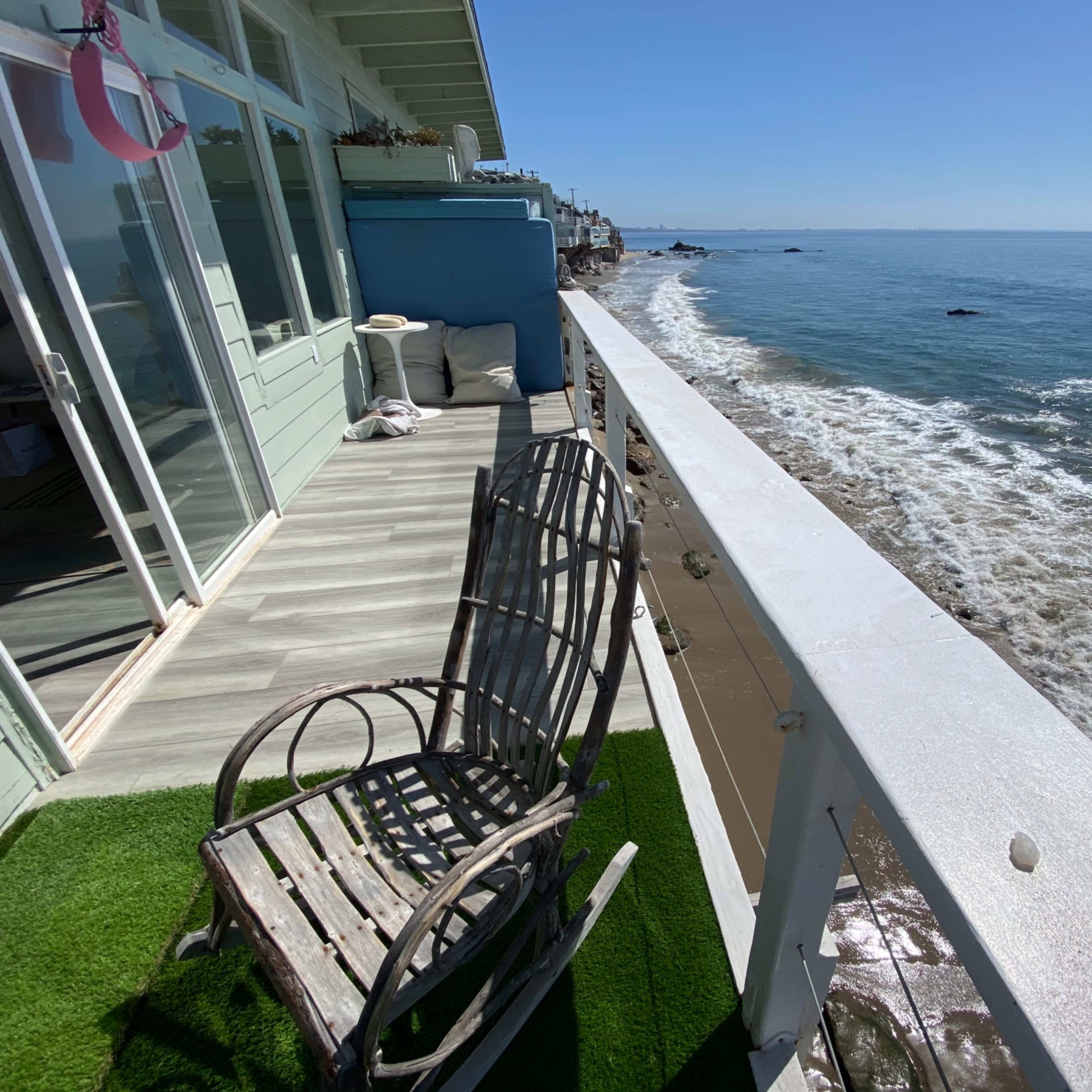 A weathered rocking chair is positioned on a balcony overlooking a sandy beach and blue ocean under a clear sky.